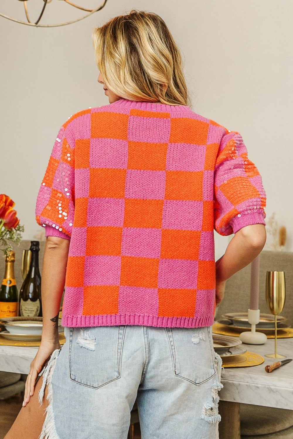 A person wearing a BiBi checkered short sleeve sequin sweater in pink and orange, paired with distressed denim shorts, stands indoors with tulips on a table in the background.