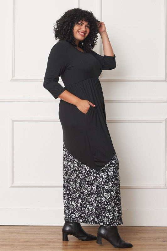 A woman with curly hair is wearing the Asymmetrical Leopard Accent Maxi Dress, featuring a long black design with a floral patterned section at the bottom. She stands against a white paneled wall, her hands tucked in her dress pockets, showcasing its elegant design.