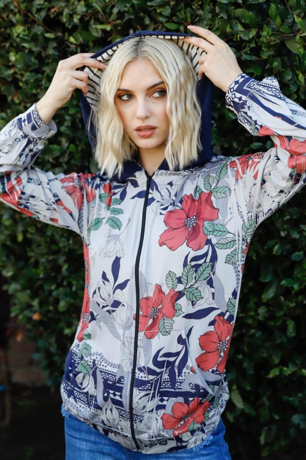 A woman stands outdoors wearing the Blumin Apparel Full Size Floral Zip-Up Hoodie with her hands in the pockets. A leafy green hedge is in the background, adding to the casual chic vibe.