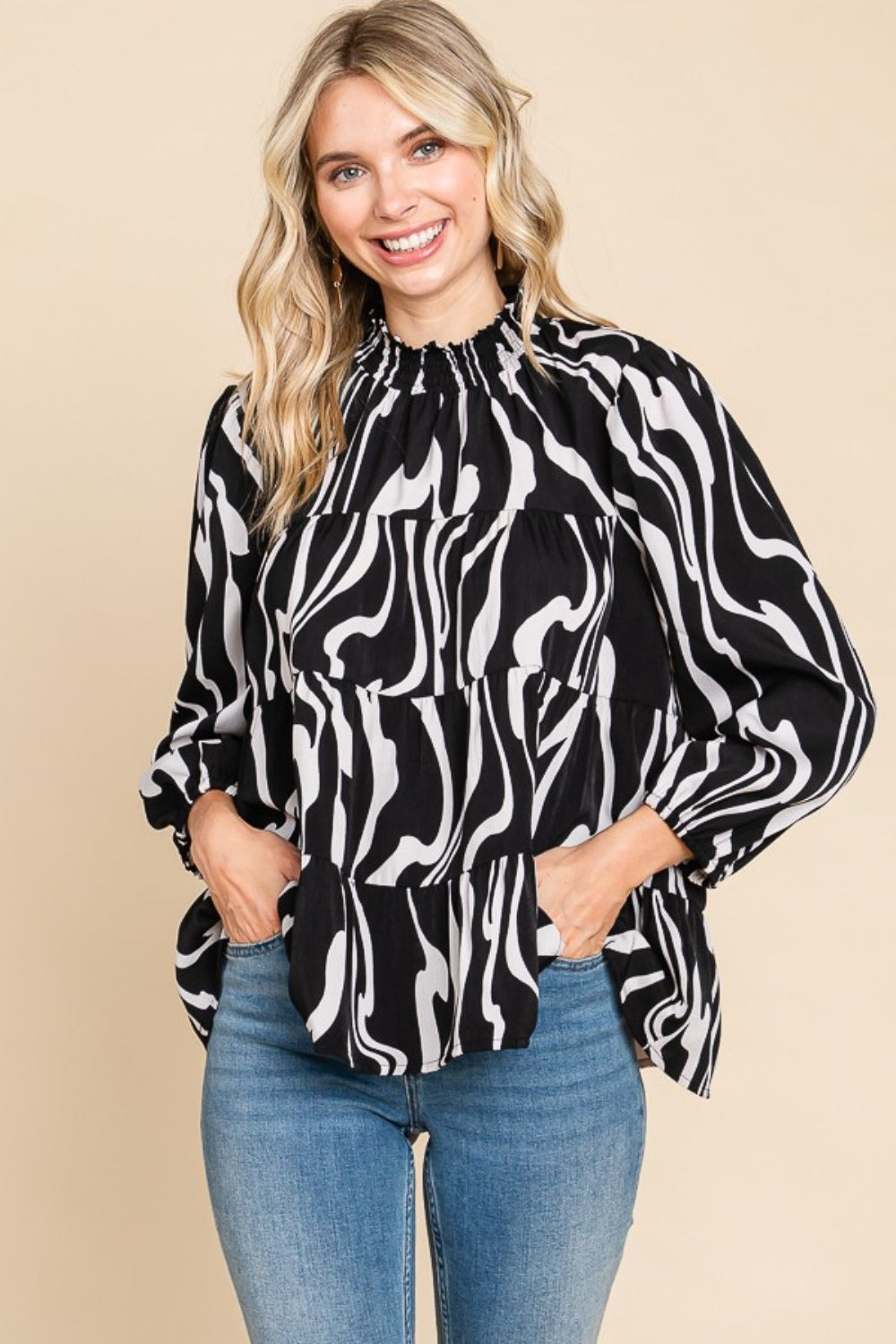 A person wearing a Culture Code Printed Smock Neck Tiered Blouse, featuring a black and white patterned design and long sleeves, paired with blue jeans, stands against a beige background.