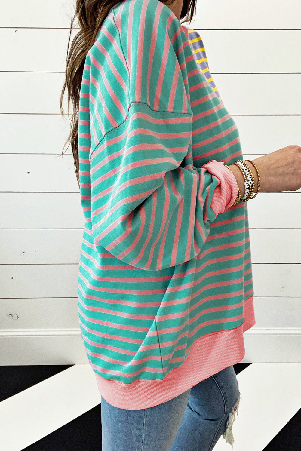 A person with wavy hair is seen from behind wearing a Casual Stripe Colorblock Drop Shoulder Oversize Sweatshirt in pastel colors and blue jeans. They are carrying a white shoulder bag, showcasing a trendy color block design.