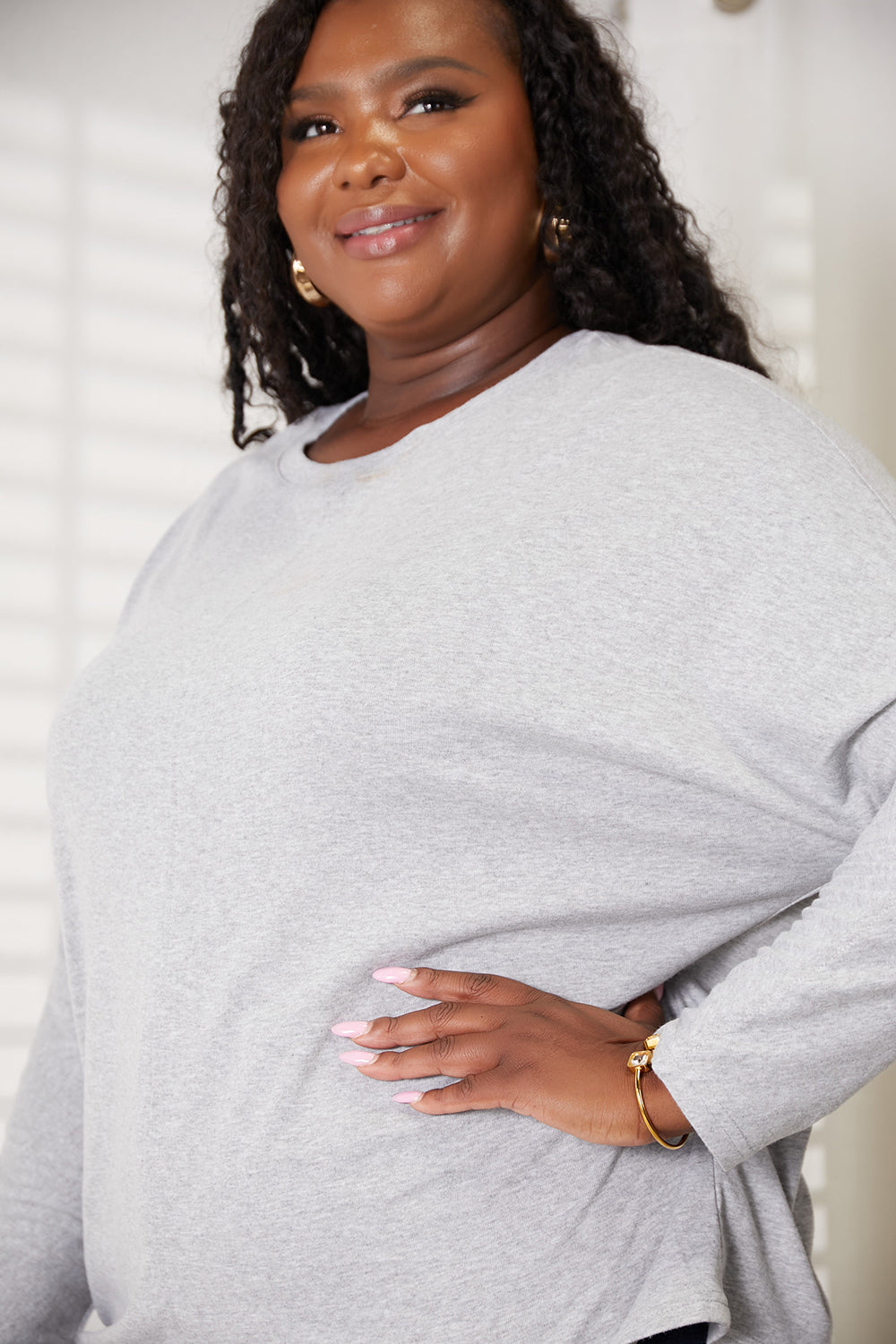 A person with long hair wearing a Double Take Seam Detail Round Neck Long Sleeve Top in solid black and blue jeans stands with one hand in their pocket, looking downward and smiling.