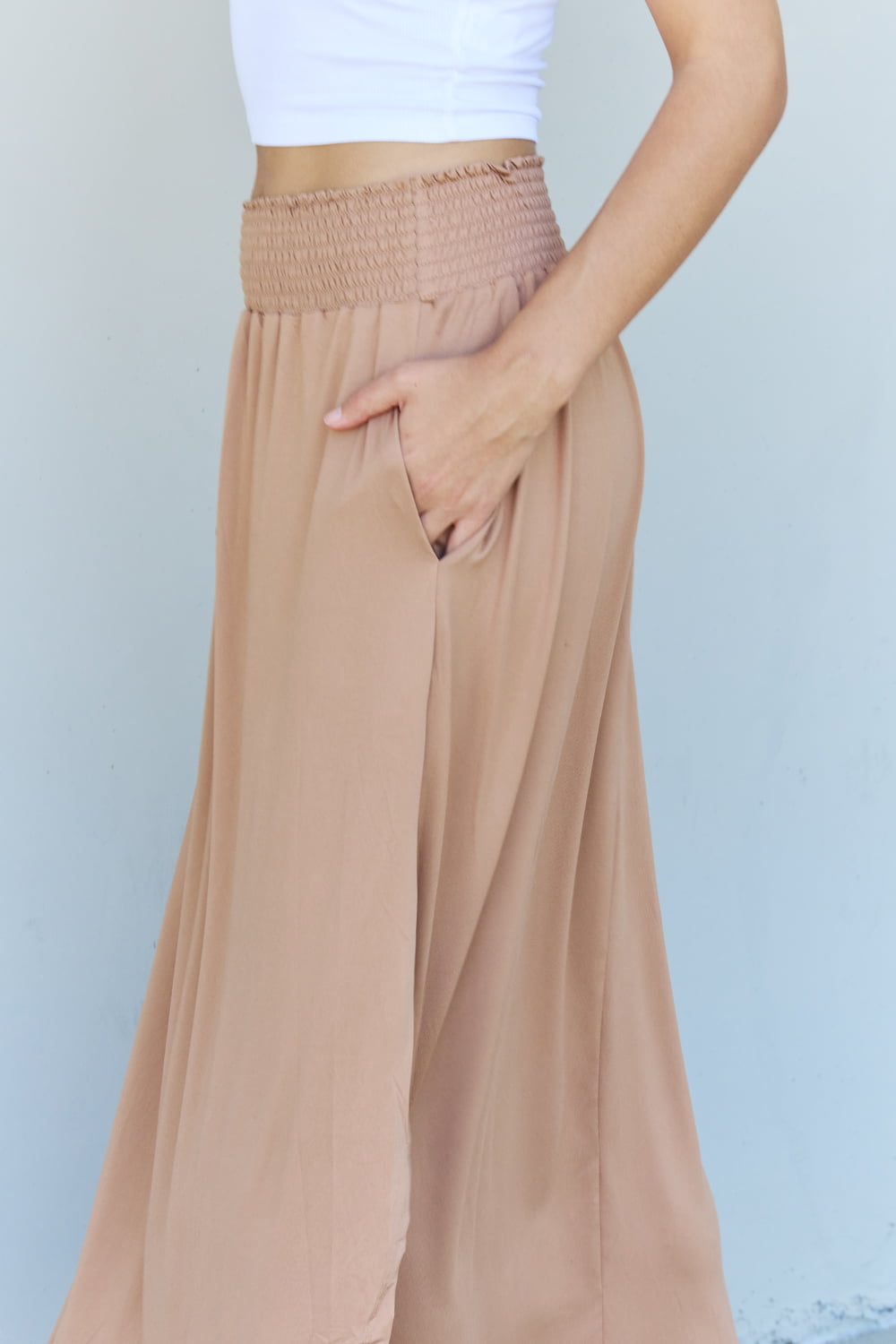 A person stands against a wall, wearing a white sleeveless top, the Doublju Comfort Princess Full Size High Waist Scoop Hem Maxi Skirt in Tan, and blue platform sandals. The right hand rests on the side of the skirt.