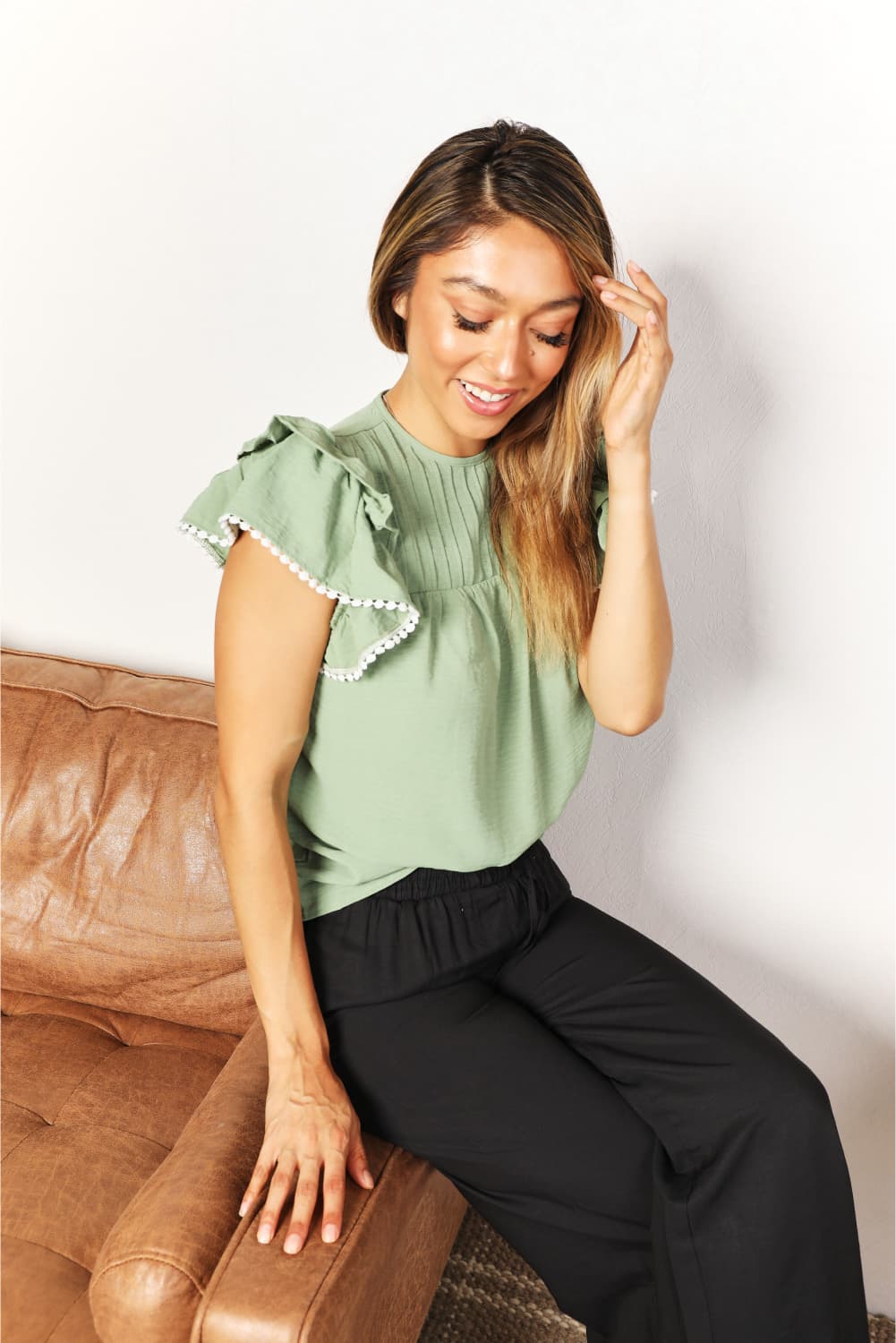 A woman stands and smiles, wearing the Perfee Pleated Detail Flutter Sleeve Blouse in light green paired with black pants.