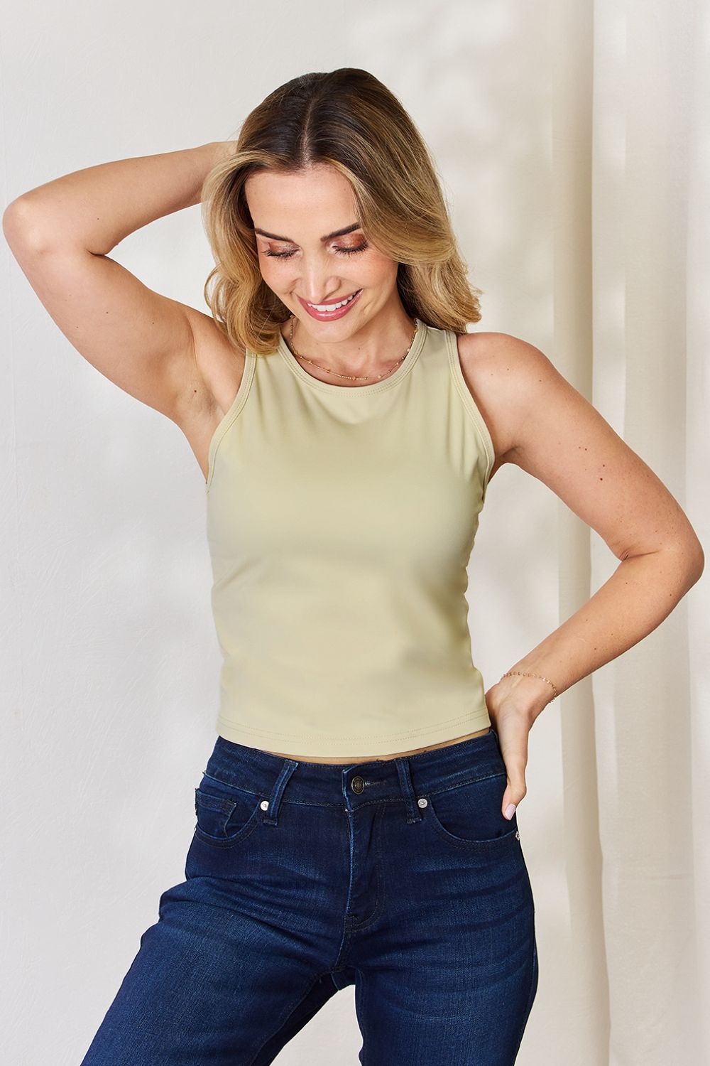 A woman with light hair wears the Basic Bae Full Size Round Neck Slim Tank in black and blue jeans, standing against a light-colored curtain.