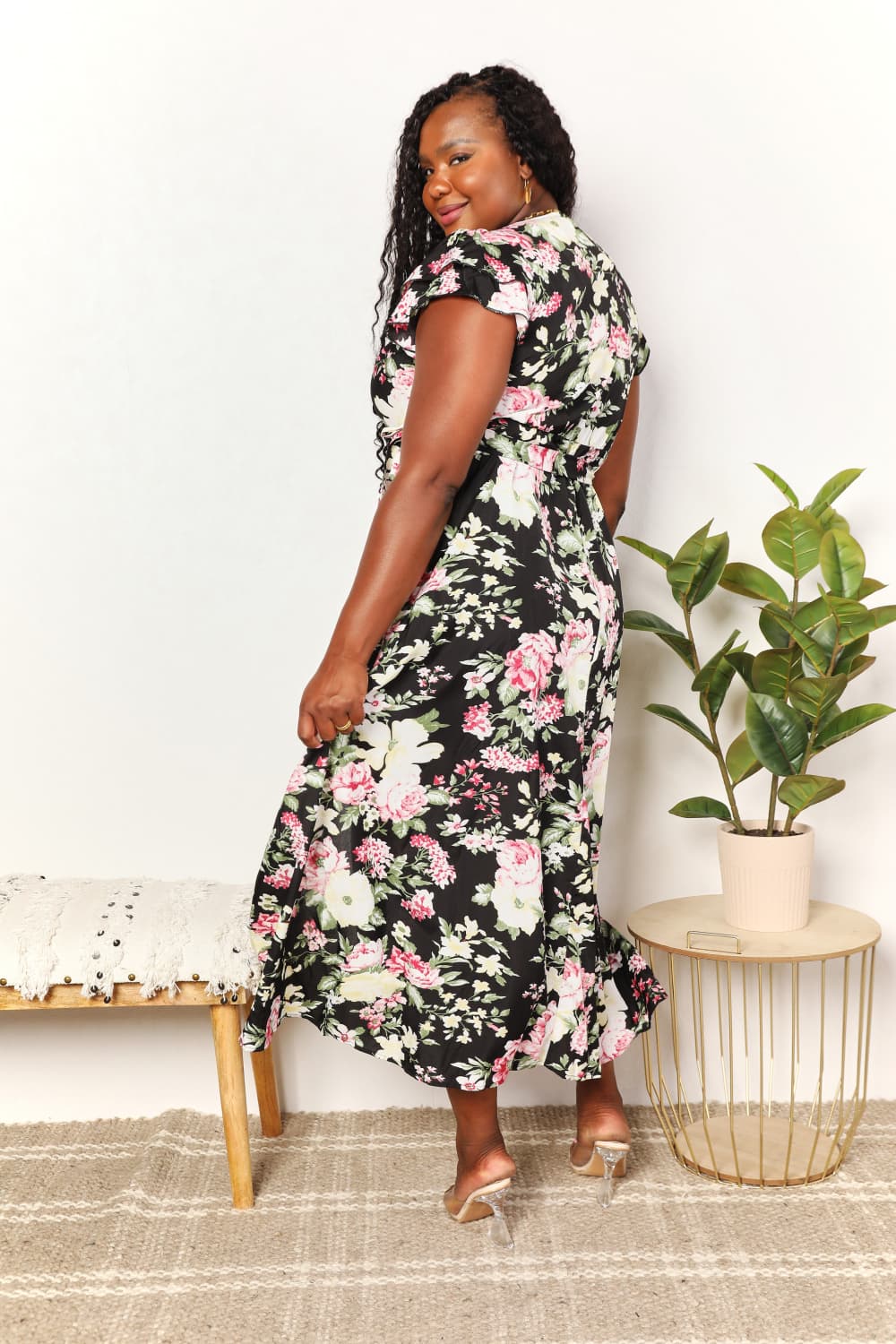 A woman wearing the Perfee Floral Flutter Sleeve Tie-Waist Split Dress stands beside a potted plant and a bench with textured fabric, perfect for adding charm to any vacation wardrobe.