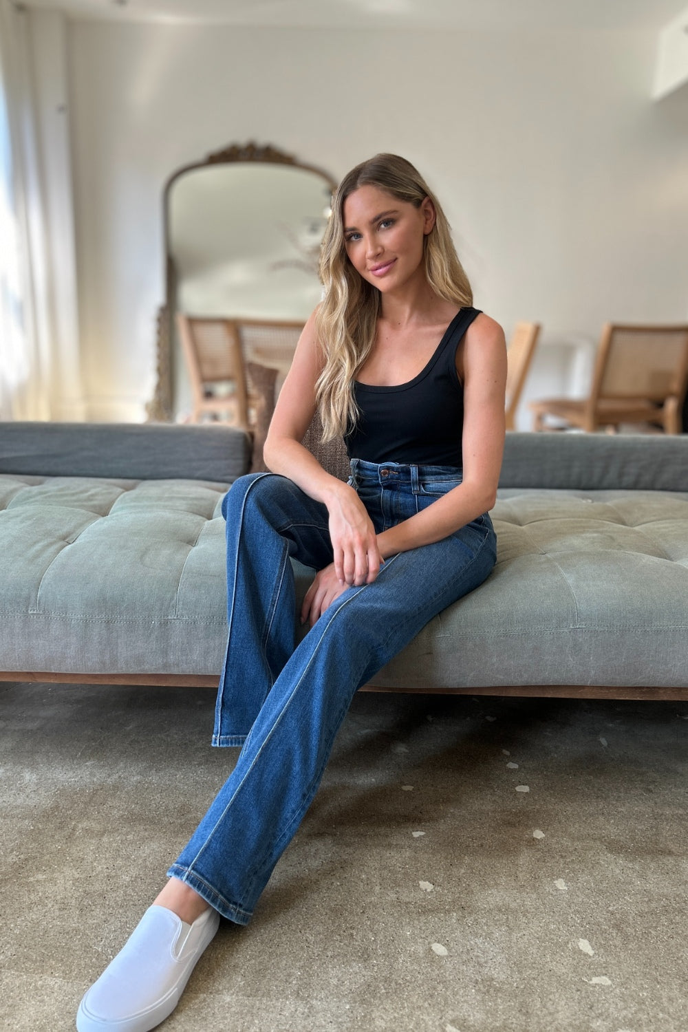 Two women stand side by side indoors, both wearing black tank tops, Judy Blue Full Size High Waist Front Seam Detail Straight Jeans, and white shoes. They are smiling and posing for the camera. A plant and shelves with decorations are in the background.