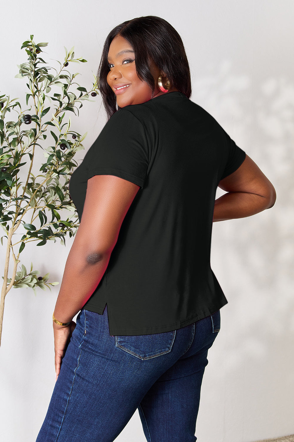 A woman with long hair, smiling with her right hand in her pocket and standing next to a plant, is wearing the Basic Bae Full Size Round Neck Short Sleeve T-Shirt in black along with beige pants made of rayon spandex.