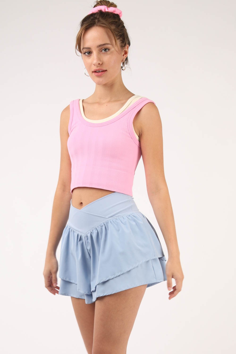 A person stands in front of a white background, wearing a pink sleeveless top and VERY J V-Shaped High Waist Layered Active Shorts in light blue. Their hair is tied with a pink scrunchie, and they are looking at the camera.