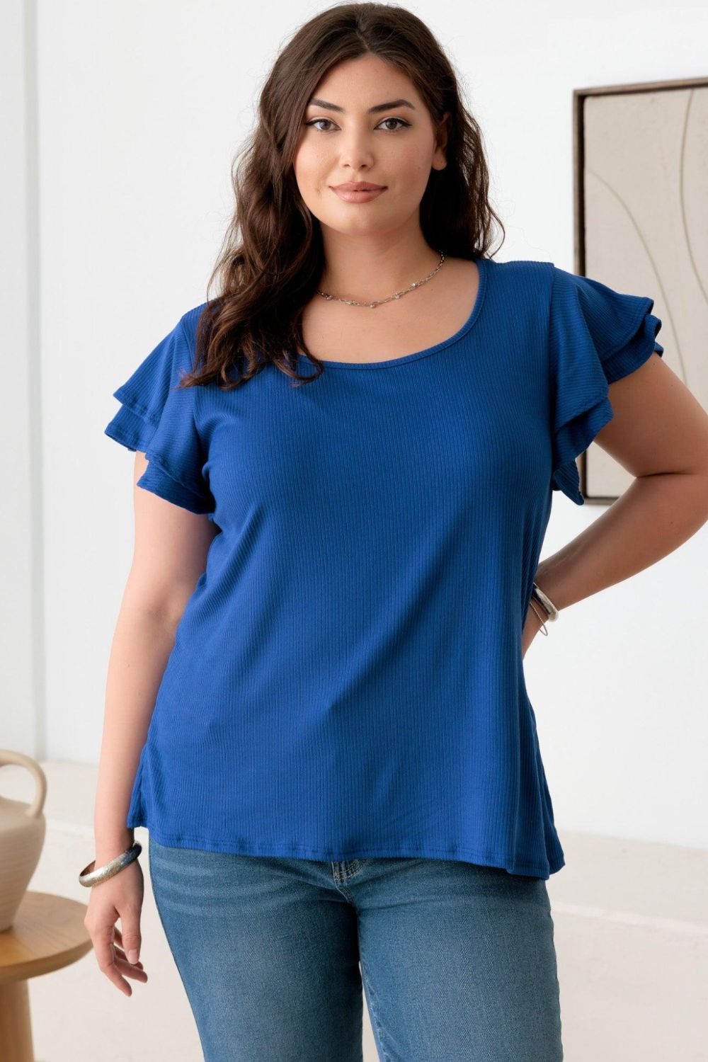 A person wearing the Gilli Plus Size Short Fluttery Sleeve Round Neck Top in blue and jeans stands indoors, showcasing an effortlessly chic style.