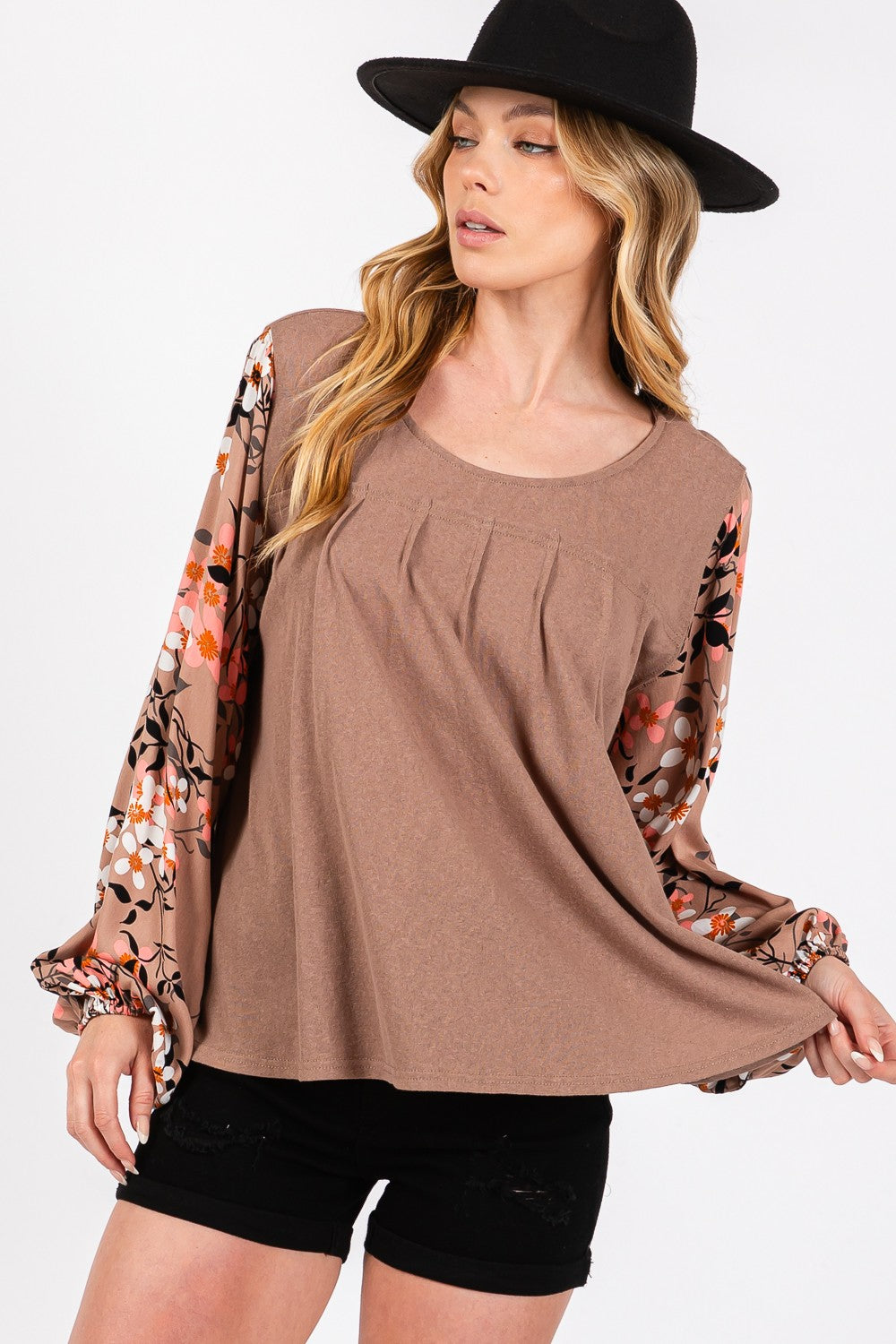 A person is wearing the SAGE + FIG Floral Long Sleeve Front Pleated Detail Blouse, paired with black shorts and a black hat, standing against a plain background.