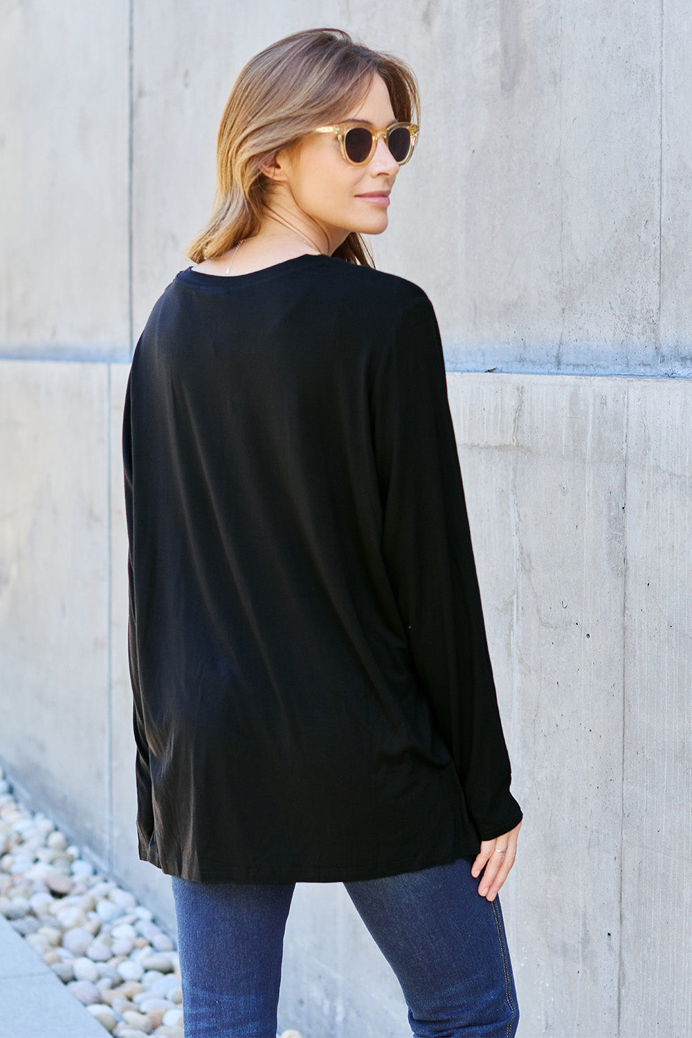 A woman with light brown hair stands against a concrete wall, wearing the Basic Bae Full Size V-Neck Long Sleeve Top in olive green and blue jeans, with a white backpack on her shoulder. Her outfit exudes basic style and is made from polyester, ensuring it’s both comfortable and suitable for machine wash cold. She is looking at the camera.