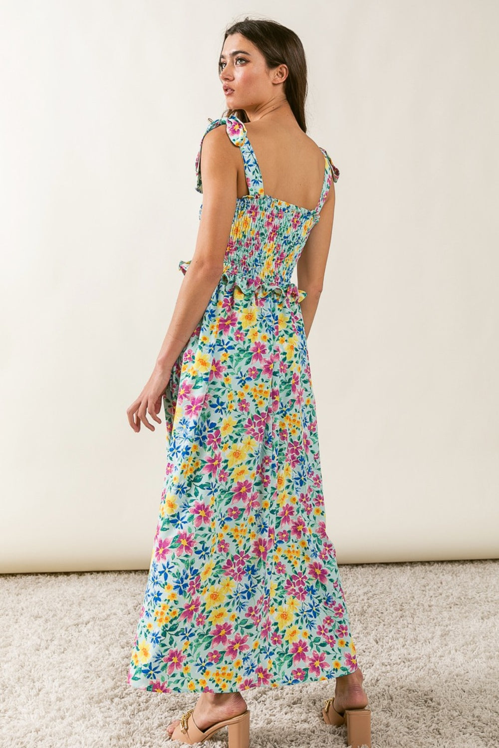 A person wearing the BiBi Floral Ruffle Trim Smocked Cami Dress with beige sandals stands on a beige carpet, posing against a white backdrop.