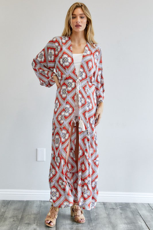 A person wearing a Printed Long Sleeve Loose Kimono with a bohemian blue and pink print over a white outfit stands in a room with a gray floor and white wall.