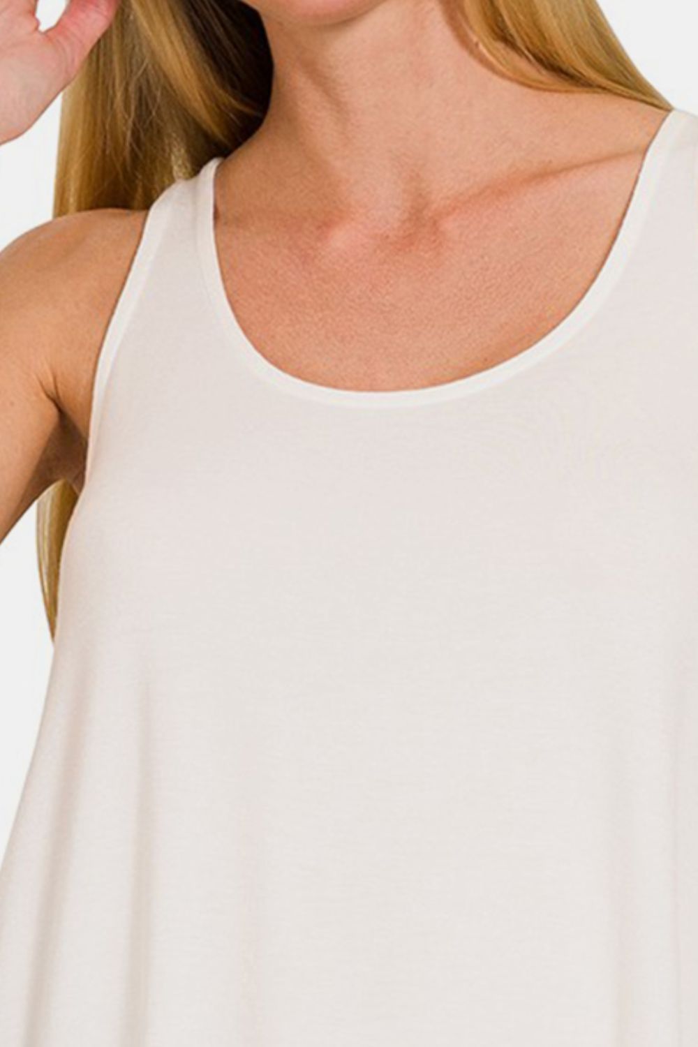 A person with long blonde hair stands against a plain background, wearing a Zenana Round Neck Flowy Hem Tank and blue jeans.