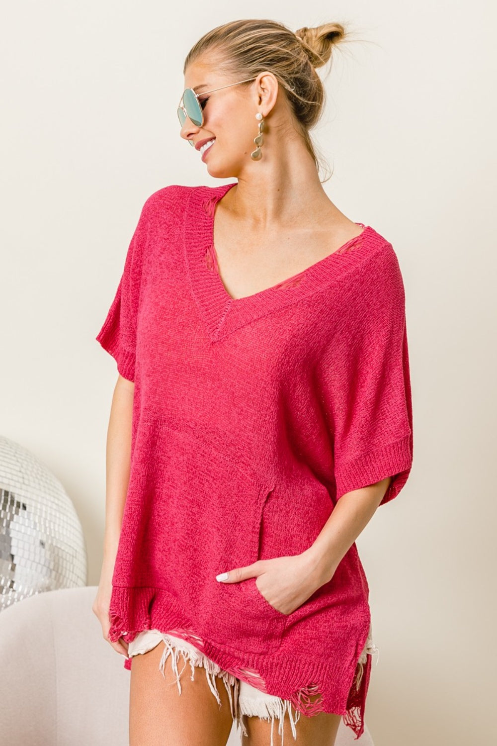 A woman in sunglasses, wearing a BiBi Distressed Hem V-Neck Slit Sweater in red and white shorts, smiles with her hands in her pockets, effortlessly showcasing her modern edge fashion.