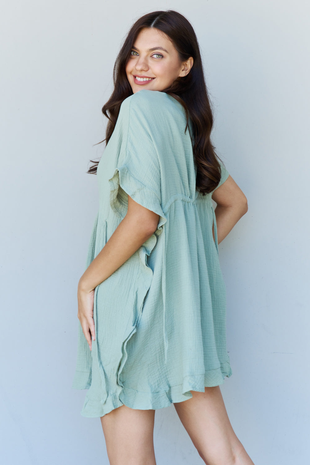 A woman is wearing the Ninexis Out Of Time Full Size Ruffle Hem Dress with Drawstring Waistband in Light Sage. Her long, dark hair falls gracefully as she smiles and looks to her left, standing against a plain background.