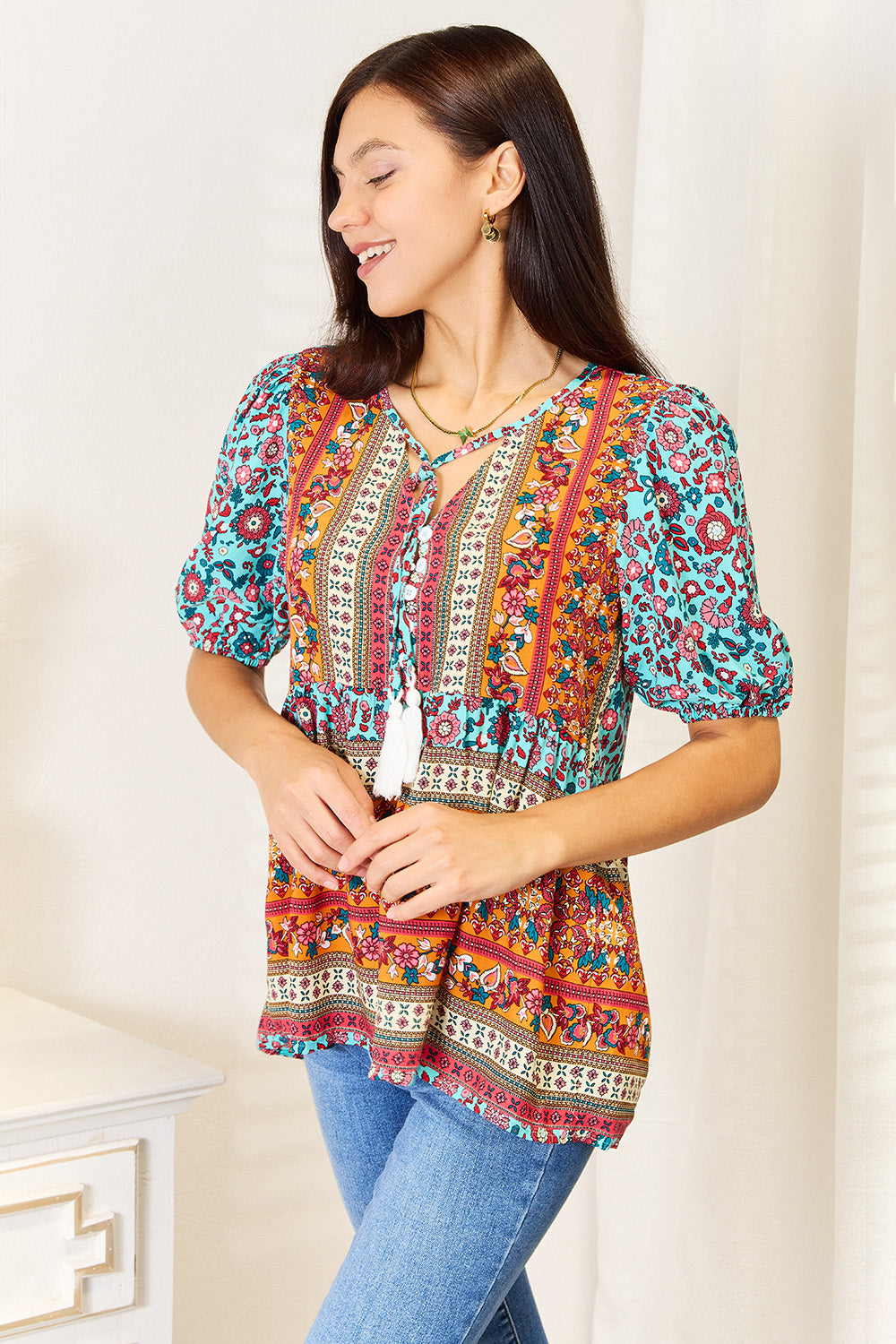 A woman wearing the Devine Bohemian Tassel Tie Puff Sleeve Babydoll Blouse, adorned with a mix of vibrant floral designs, smiles in her jeans while standing indoors against a light background.