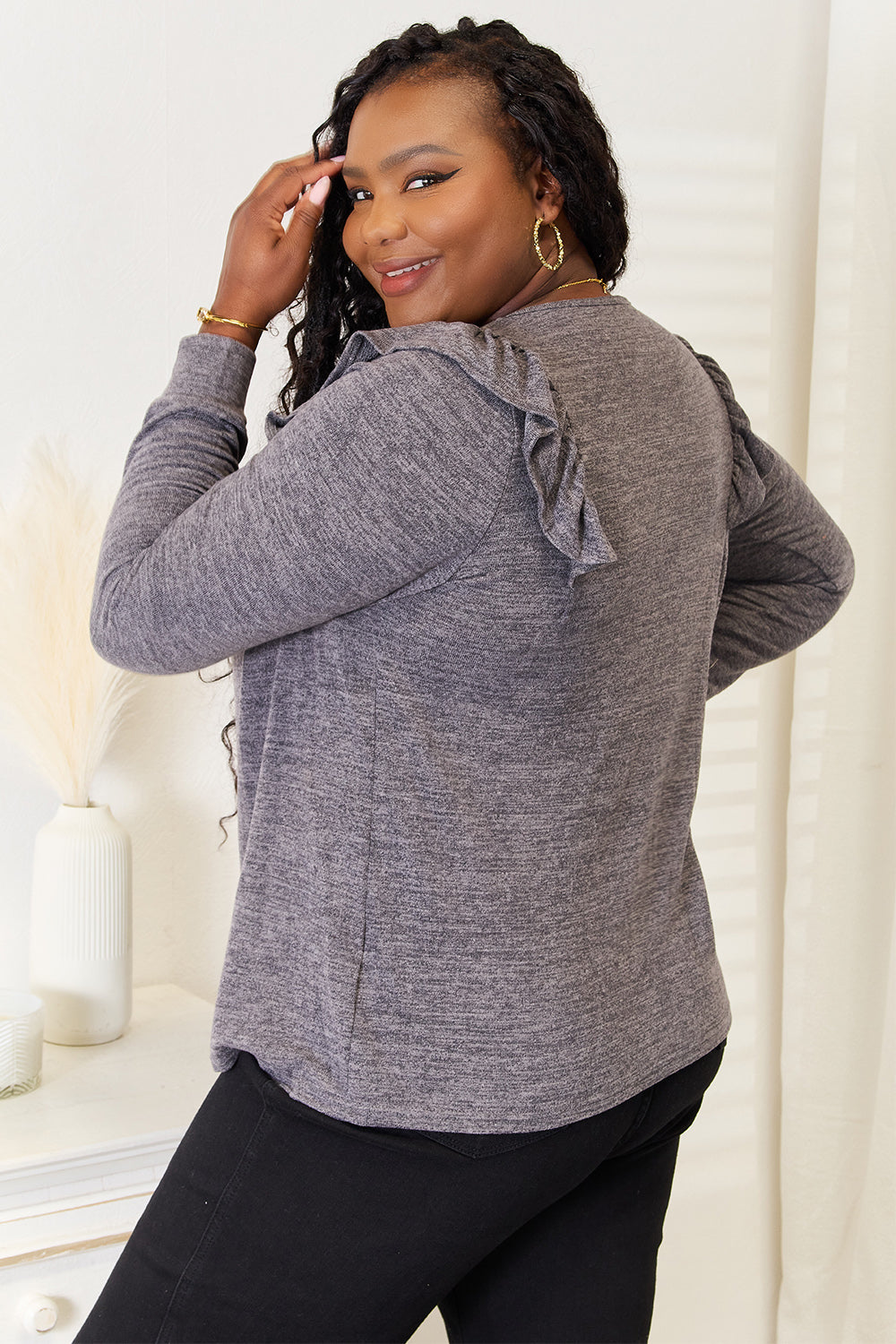 A woman wearing a Double Take Square Neck Ruffle Shoulder Long Sleeve T-Shirt in pink and blue jeans stands with one hand in her pocket, smiling. She has long curly hair and wears a necklace and bracelet. She stands in a room with white decor.