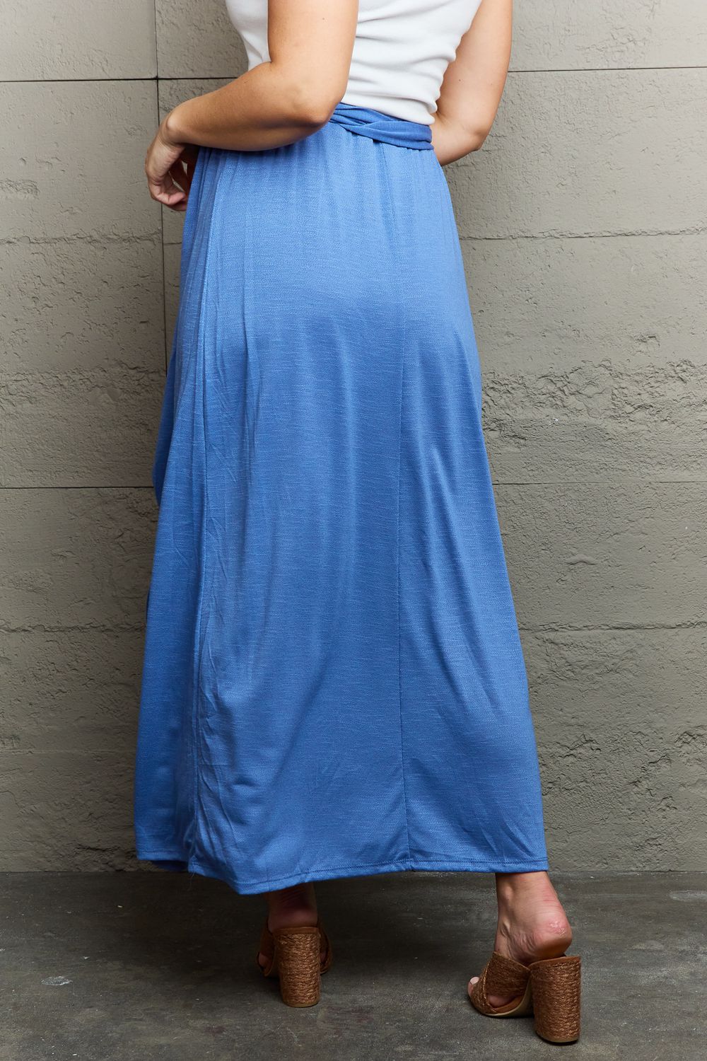 A person stands against a textured gray wall, smiling in the Ninexis Know Your Worth Criss Cross Halter Neck Maxi Dress in blue, featuring a stylish cut-out design. One hand rests on the wall while the other is confidently placed on their hip.