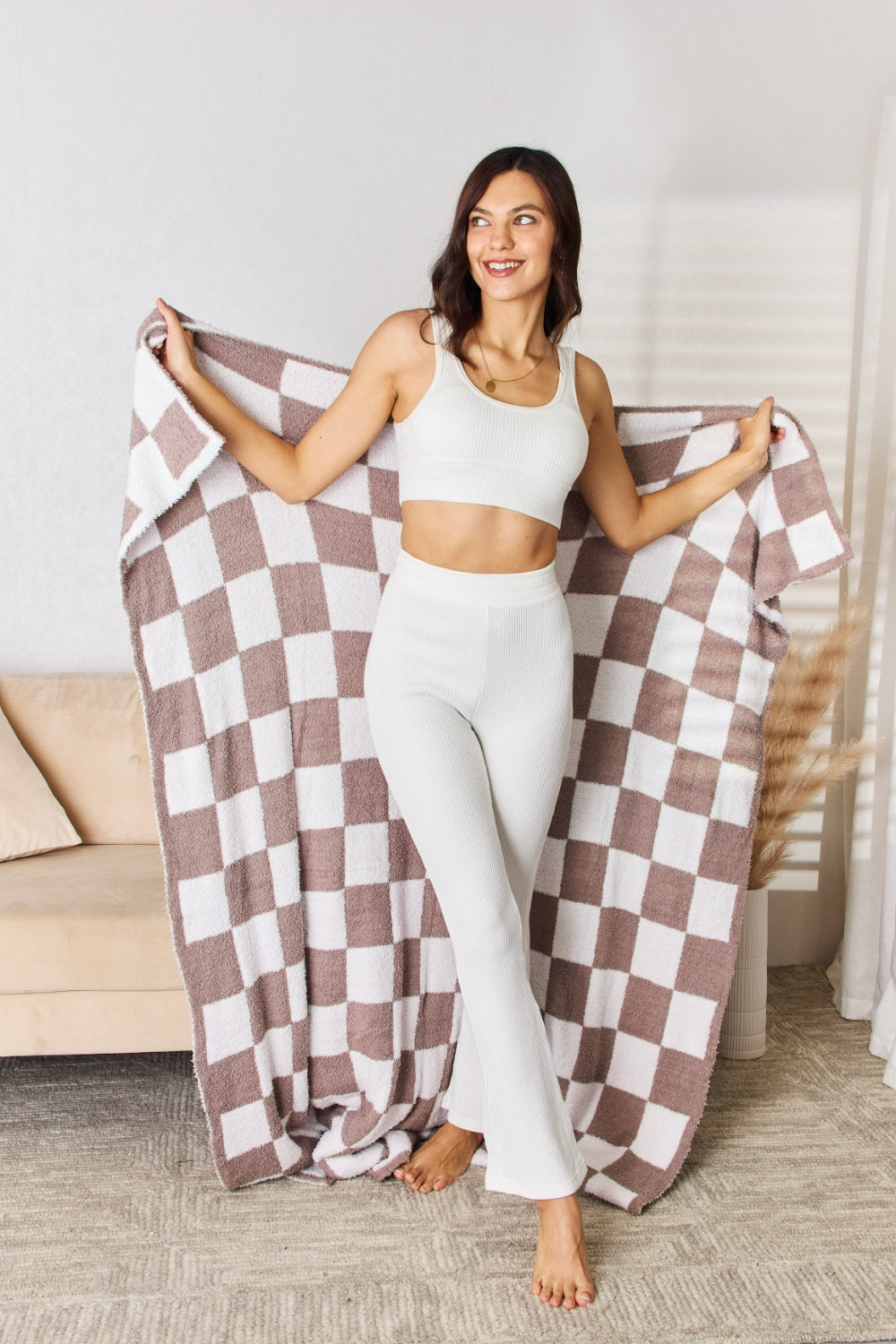 A person stands holding a large Cuddley Checkered Decorative Throw Blanket, smiling, in a room with a sofa and a potted plant. The blanket features a black and white checkerboard pattern, is made from 100% polyester, and imported for quality.