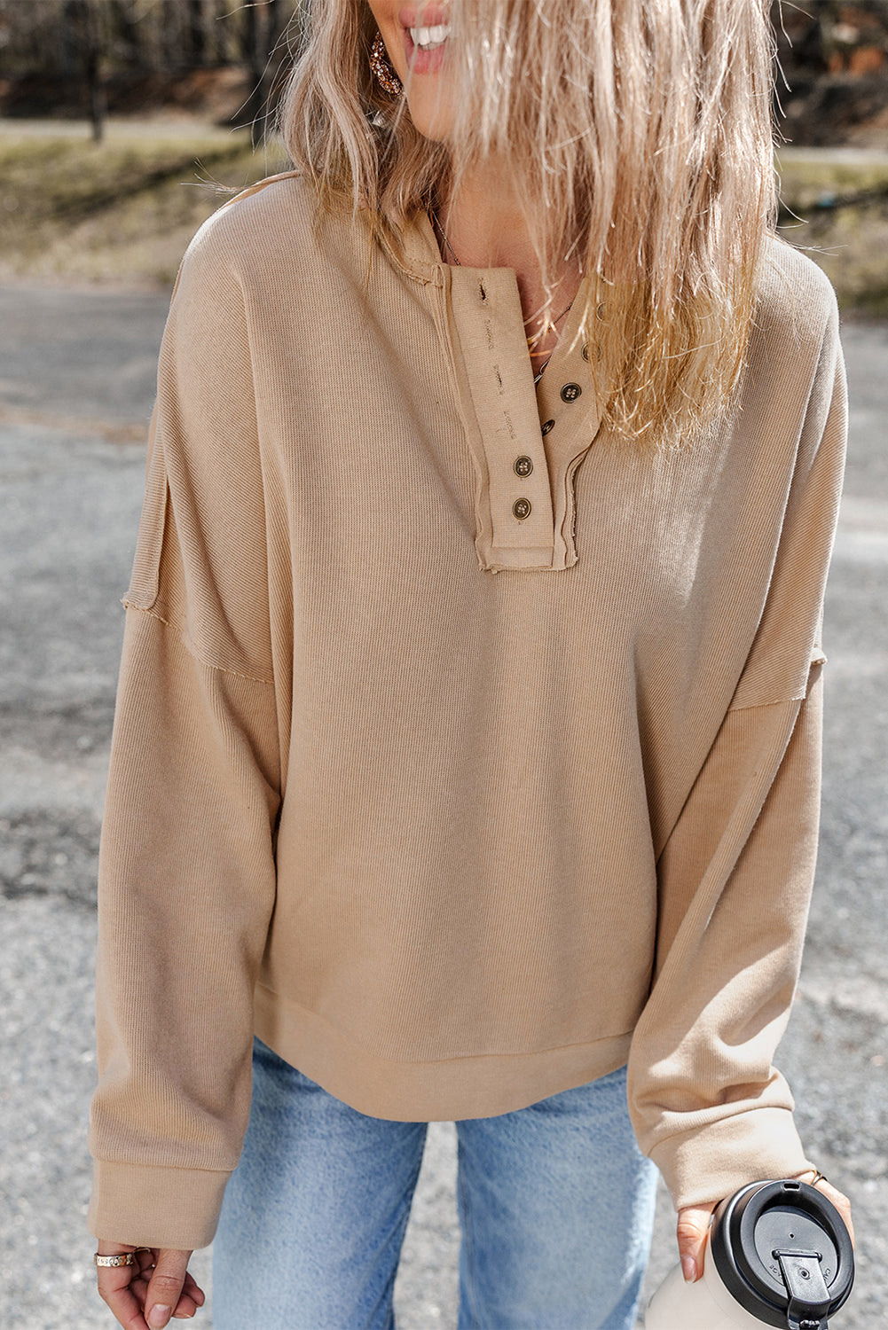 Person wearing a Khaki Casual Button Solid Patchwork Trim Hoodie and blue jeans is standing outdoors holding a white tumbler. The individual's blonde hair is partially visible from behind.