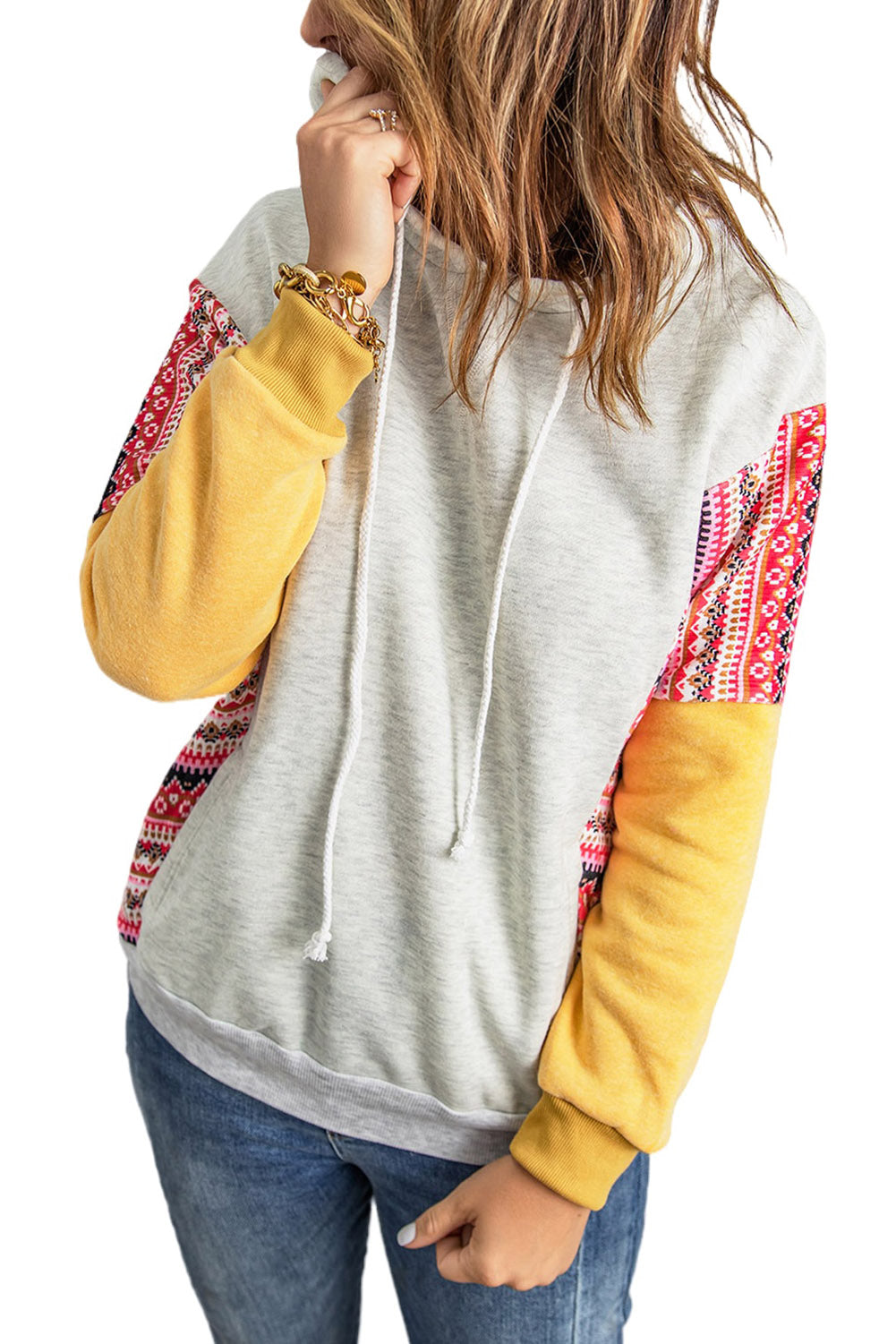 A person with long hair wearing an Aztec Patch Pullover Hoodie and blue jeans is facing away from the camera against a textured gray wall.