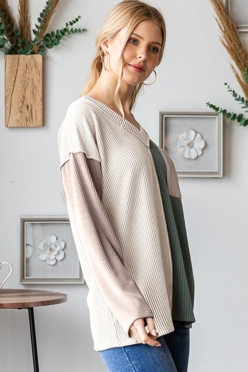 A woman stands indoors, wearing a Heimish Color Block Exposed Seam Ribbed T-Shirt with beige and green sections, paired with blue jeans. She has long blonde hair tied in a ponytail and is smiling slightly.