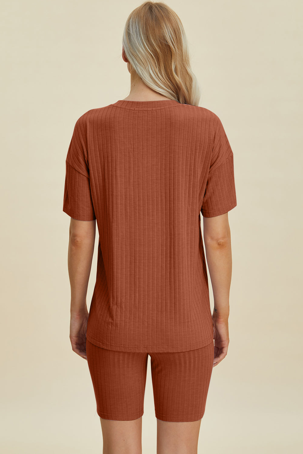 A woman with long blonde hair wearing the Basic Bae Full Size Ribbed V-Neck Short Sleeve Top and Shorts Set in rust stands against a plain background. This stylish two-piece set is made from a comfortable, stretchy fabric that is also machine washable.