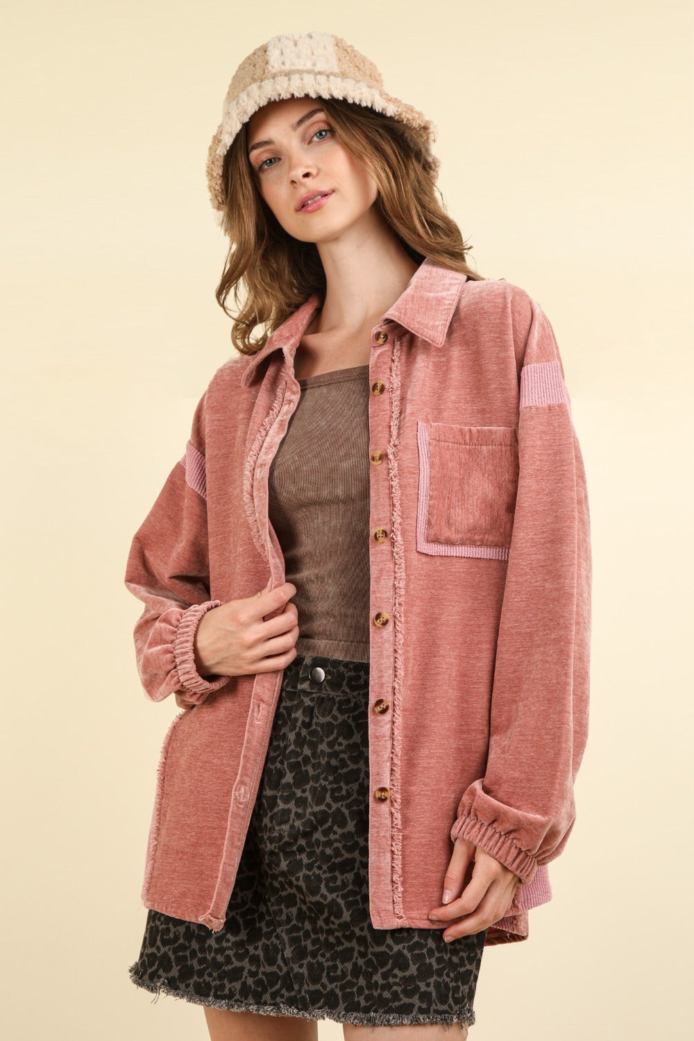 A person showcasing the VERY J Mixed Media Button Down Raw Hem Shacket in pink, paired with a black patterned skirt with a raw hem and a beige hat, stands against a light background.