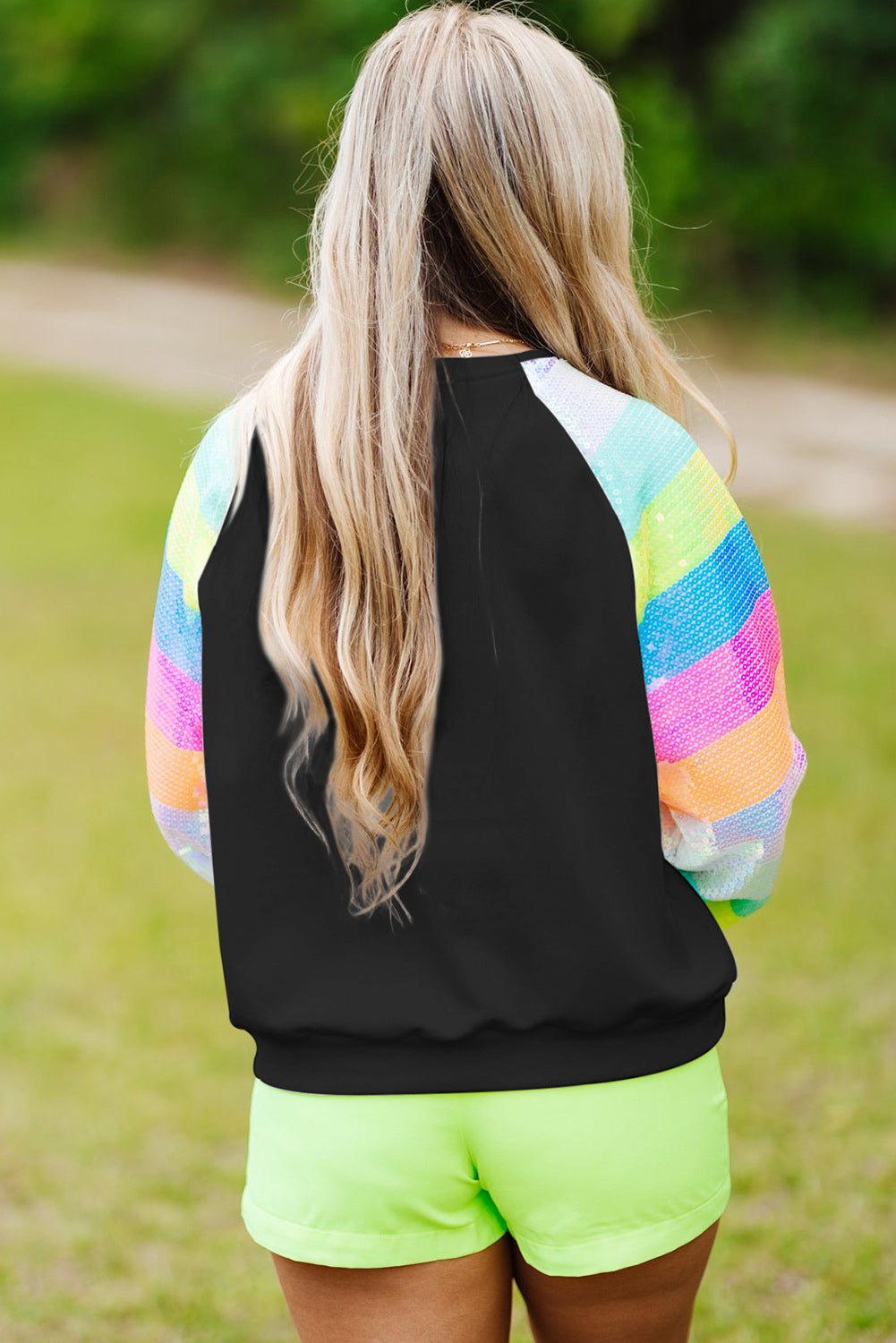 A person with shoulder-length hair is wearing a Black Sequin Color Block Raglan Sleeve Pullover Sweatshirt and blue jeans. They are facing away from the camera in a room with a potted plant.