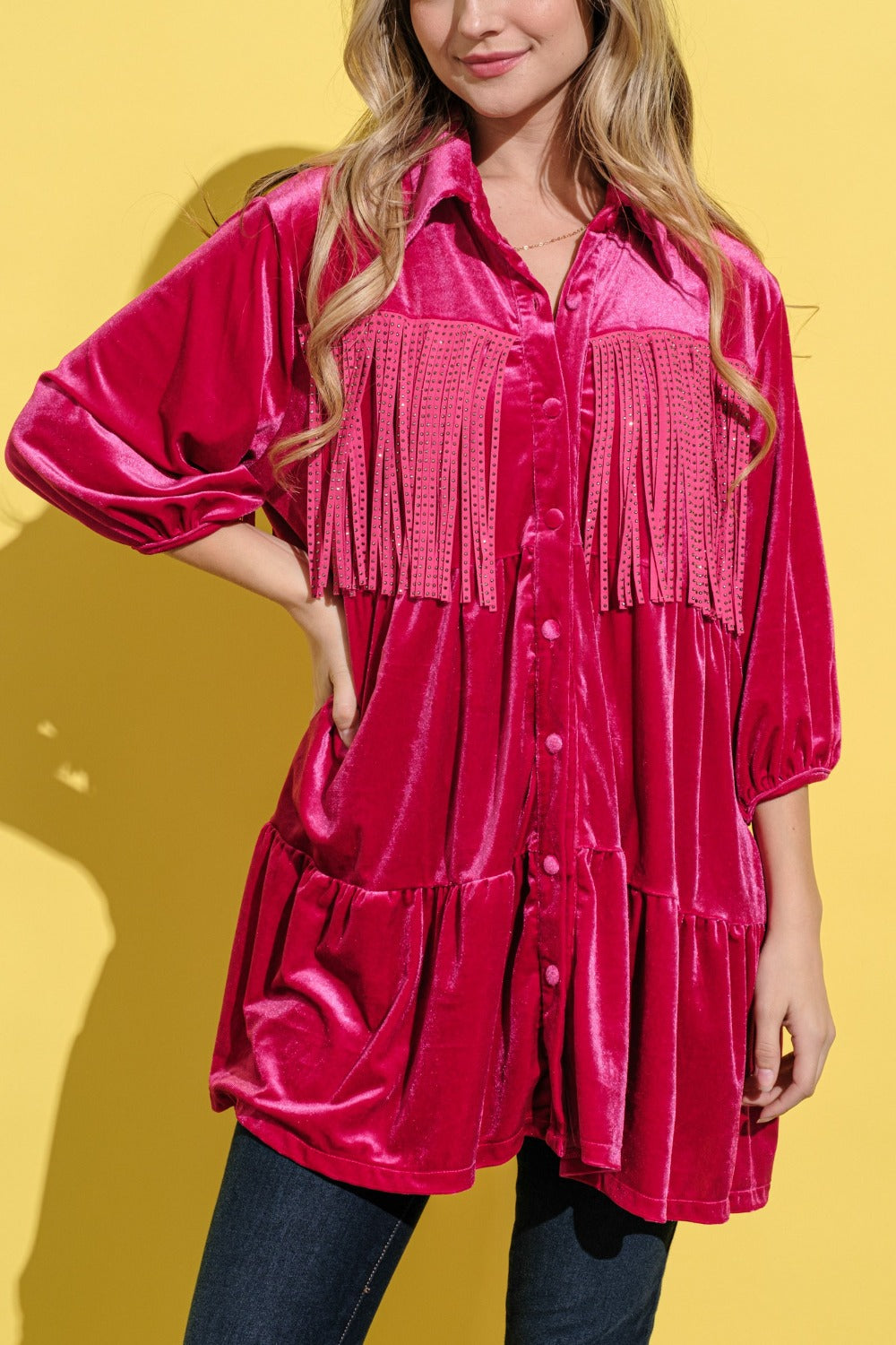 A woman stands against a bright yellow background, smiling and wearing an elegant statement fringed, bright pink And The Why Fringe Detailed Velvet Shirt Dress.