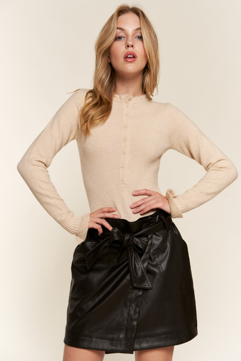 A woman in a beige sweater and black leather skirt stands against a neutral background, her look perfectly complemented by the sleek lines of the And The Why Half Button Round Neck Long Sleeve Bodysuit underneath.