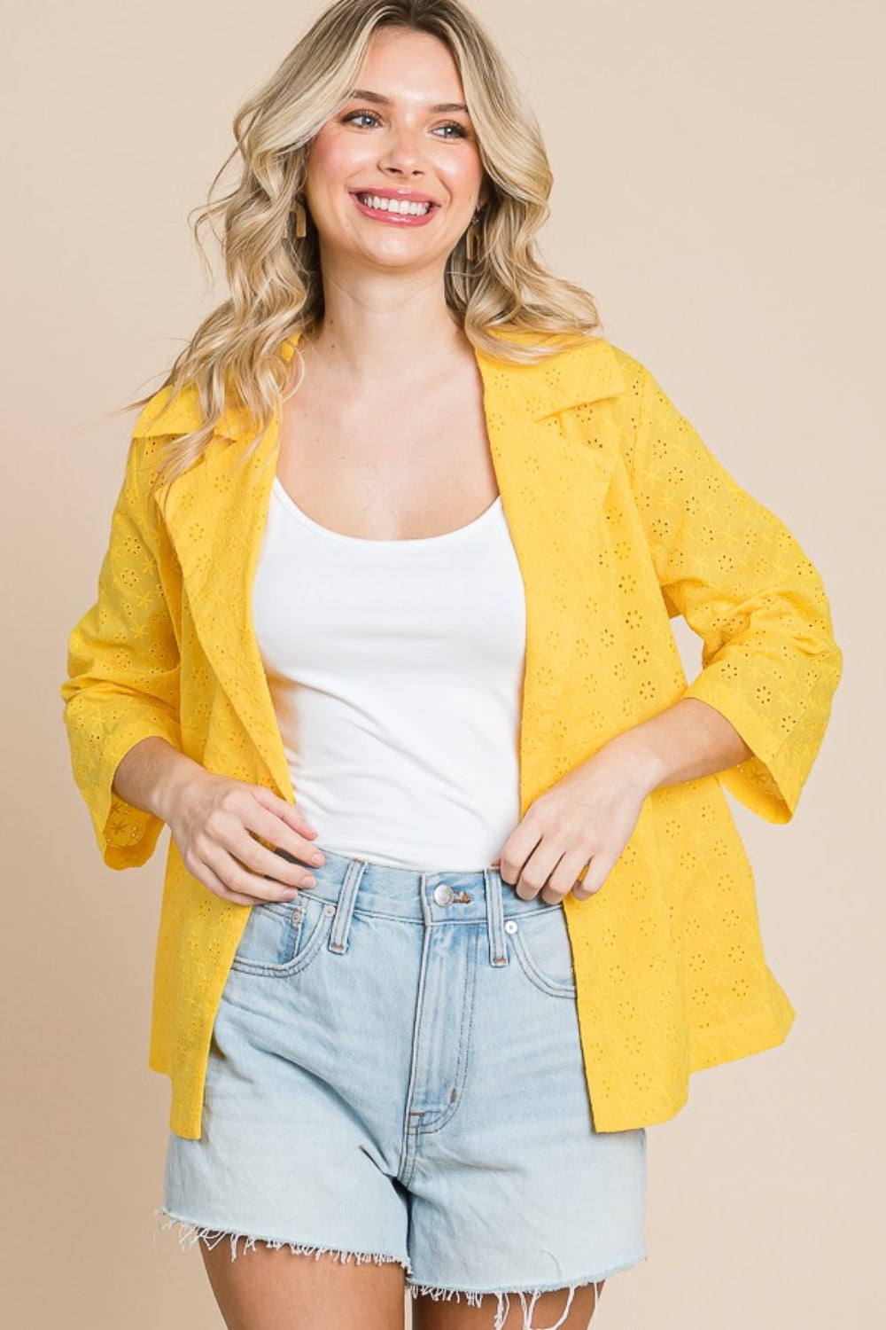 Woman in a white tank top, denim shorts, and the Culture Code Double Breasted Eyelet Jacket with Pockets in yellow, smiling against a beige background.