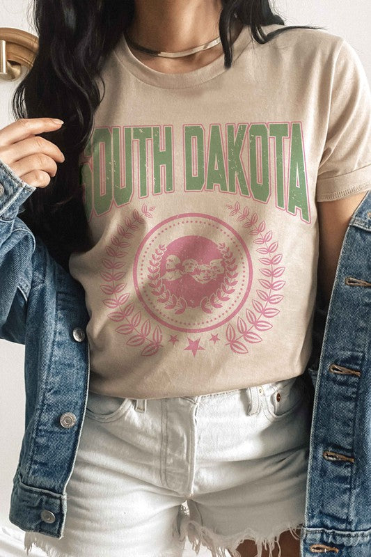 A person wearing a white SOUTH DAKOTA Graphic Tee, adorned with green and pink lettering, along with matching white shorts, stands indoors. The unisex sizing 100% cotton tee adds a comfortable yet stylish touch to their outfit.