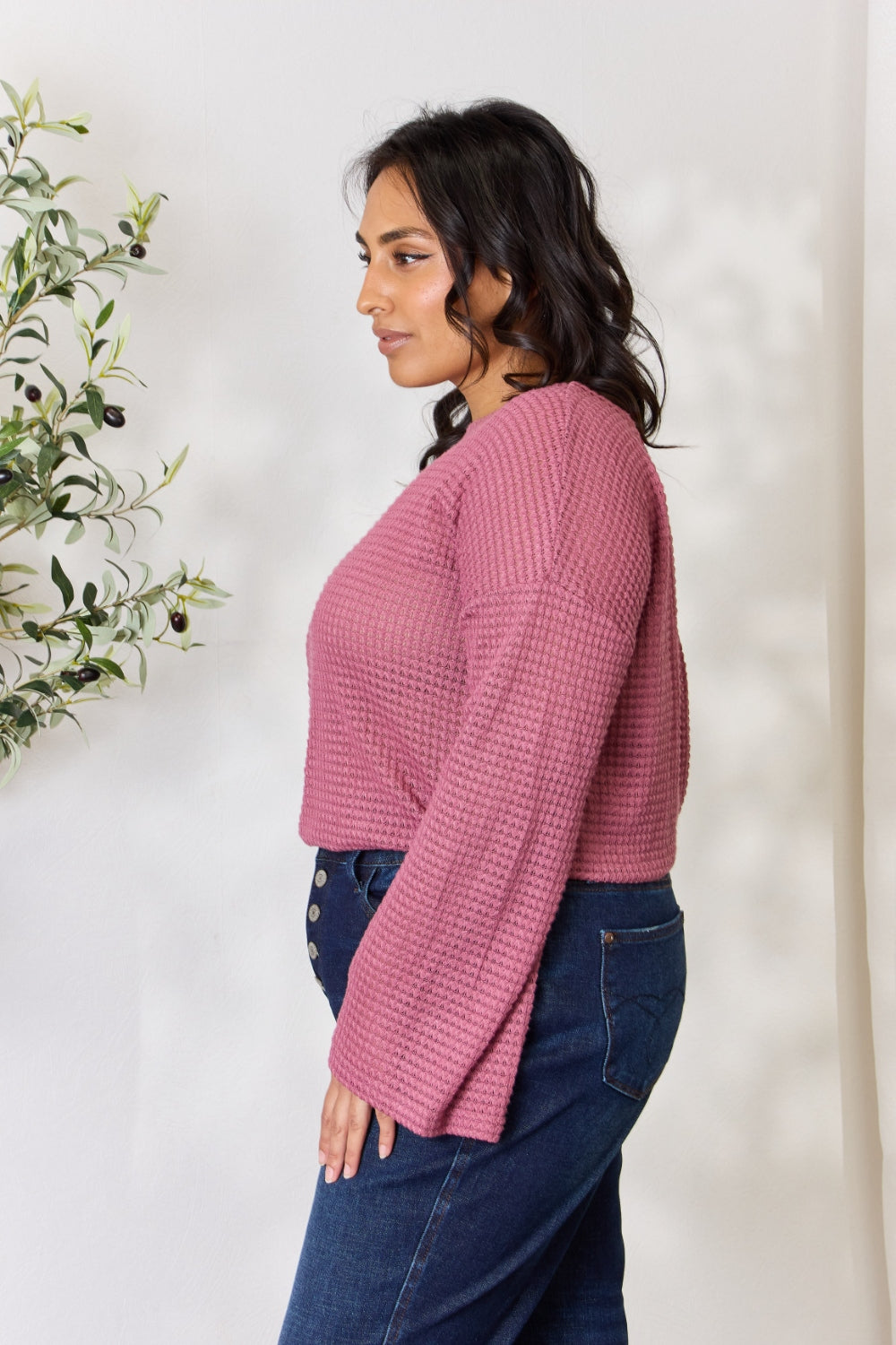 A person wearing a Culture Code Full Size Waffle-Knit Round Neck Long Sleeve Blouse in pink and jeans stands in front of a light background, complemented by a leafy plant on the left.