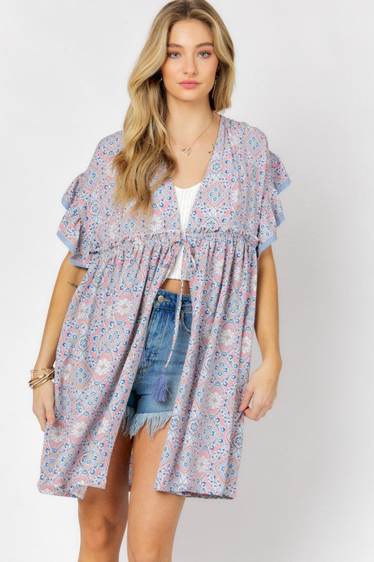 A woman stands against a white background wearing the Printed Short Sleeve Ruffle Kimono over a white top and denim shorts, effortlessly blending style and comfort.