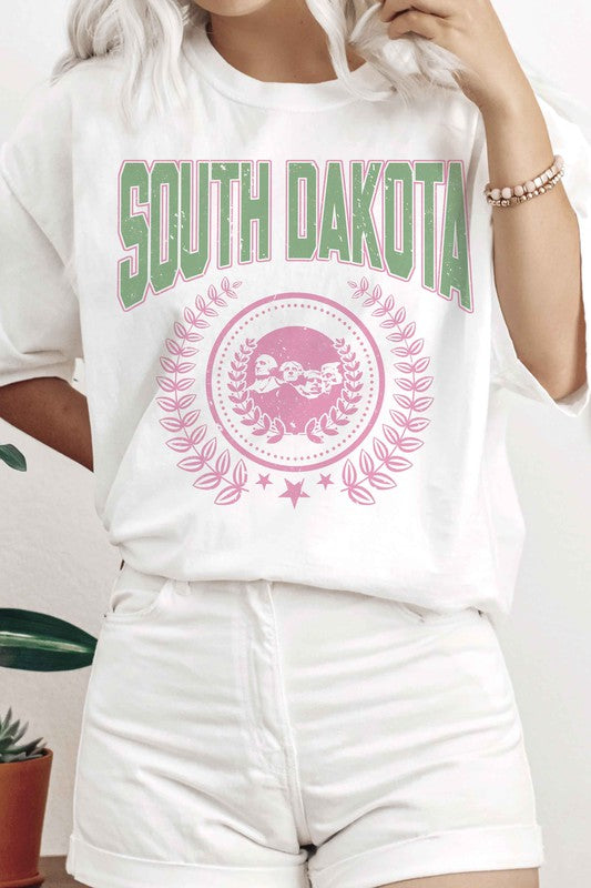 A person wearing a white SOUTH DAKOTA Graphic Tee, adorned with green and pink lettering, along with matching white shorts, stands indoors. The unisex sizing 100% cotton tee adds a comfortable yet stylish touch to their outfit.