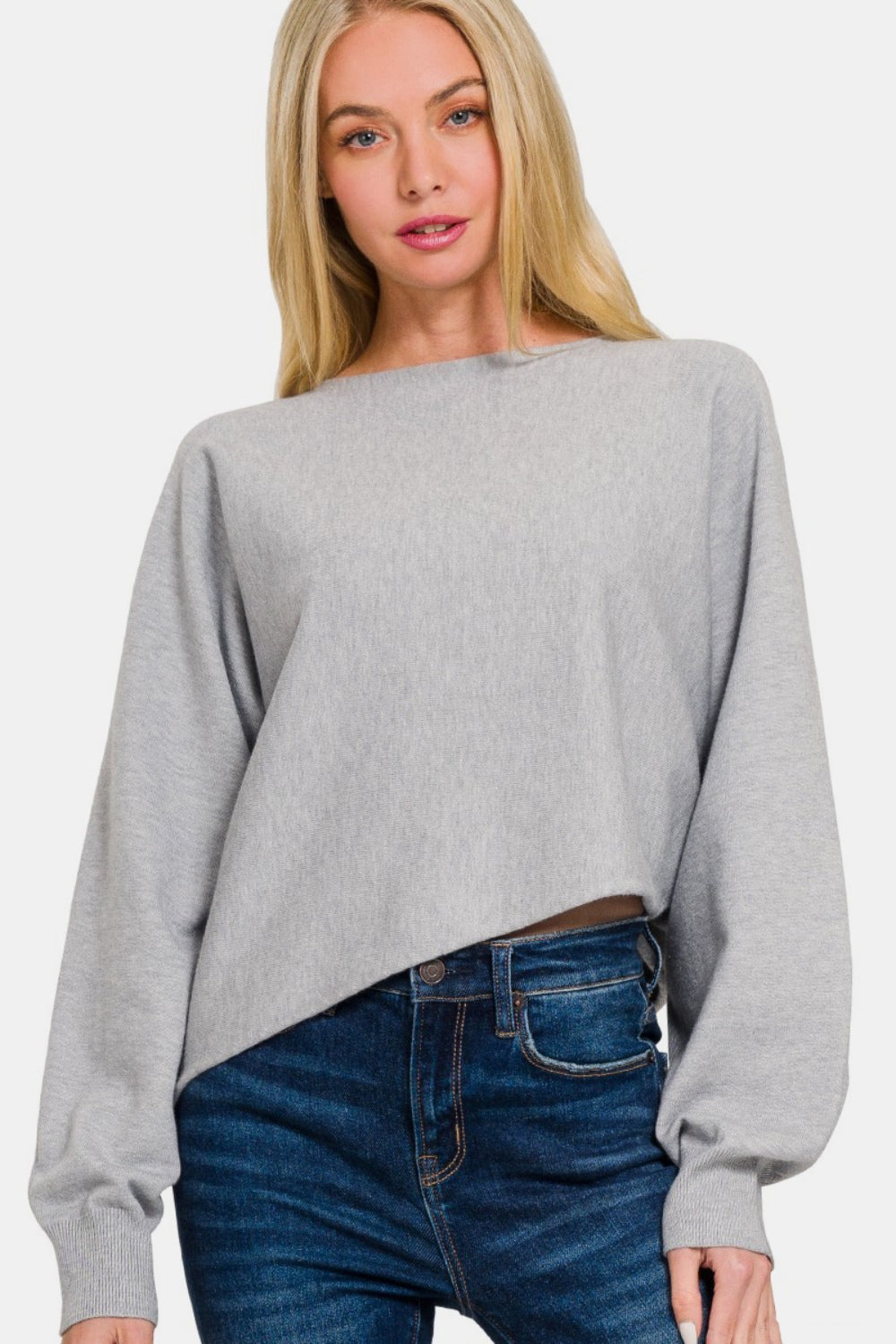 A person with long blonde hair wears a stylish Zenana Asymmetric Hem Long Sleeve Sweater made of soft and comfortable gray fabric, paired with blue jeans, standing against a plain white background.