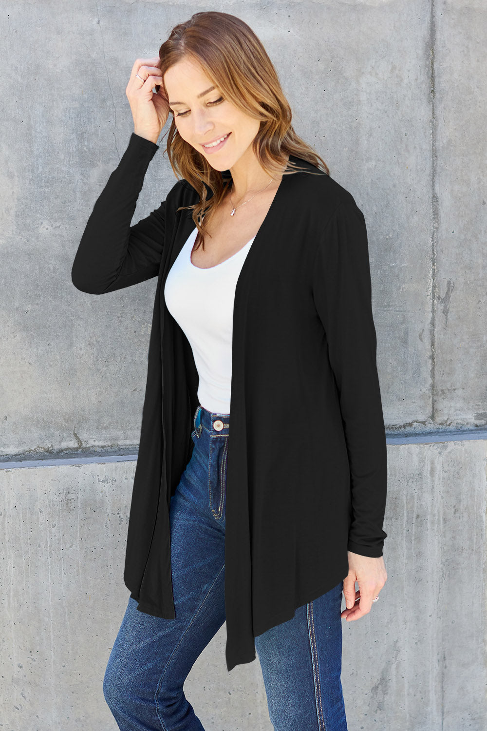 A woman wearing the Basic Bae Full Size Open Front Long Sleeve Cardigan in navy blue, paired with a white top and blue jeans, stands against a concrete wall. She touches her hair and gazes to the side, embodying a chic yet simple style. The slightly stretchy materials of the cardigan ensure comfort while maintaining its stylish simplicity. For easy care, machine wash it cold.