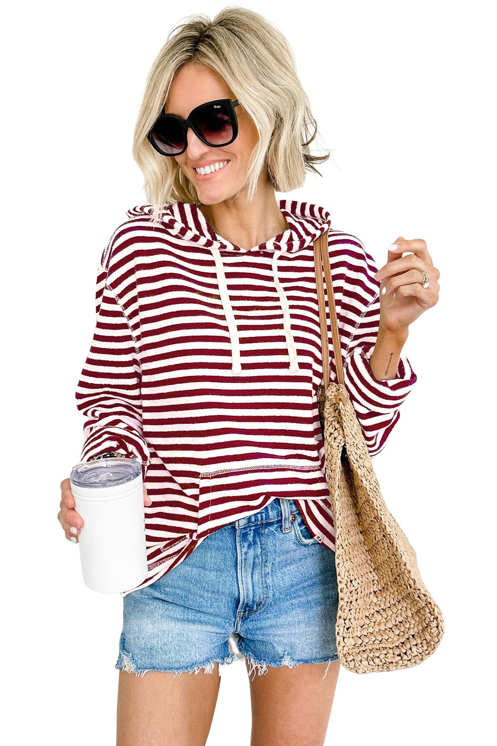 A person wearing a trendy Burgundy Stripe Kangaroo Pocket Drawstring Hoodie, paired with denim shorts and sunglasses, holds a white tumbler and a woven bag in front of a white paneled wall.