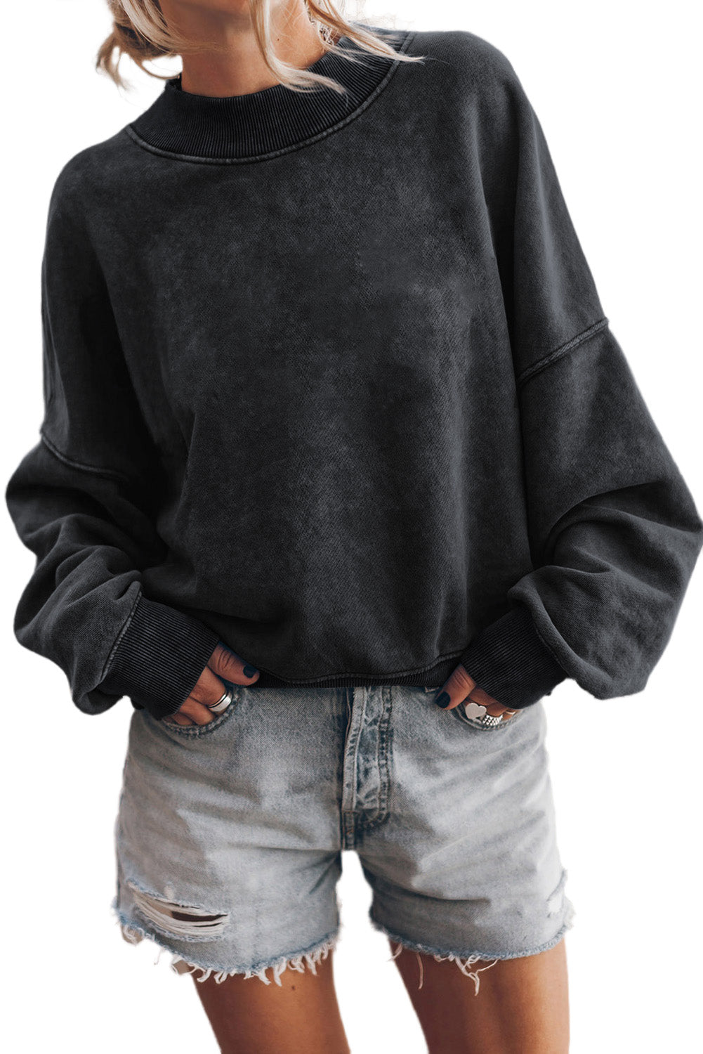 Person wearing a black drop shoulder crew neck pullover sweatshirt with rolled-up sleeves and light blue jeans, standing with one hand in their pocket, showing multiple bracelets on their wrist.