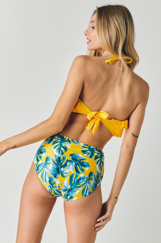 A person wearing the Solid Ruffle Top And Printed Bottom Swimsuit, featuring a vibrant yellow ruffle top and high-waisted bottoms with a blue leaf pattern, poses gracefully against a plain background.