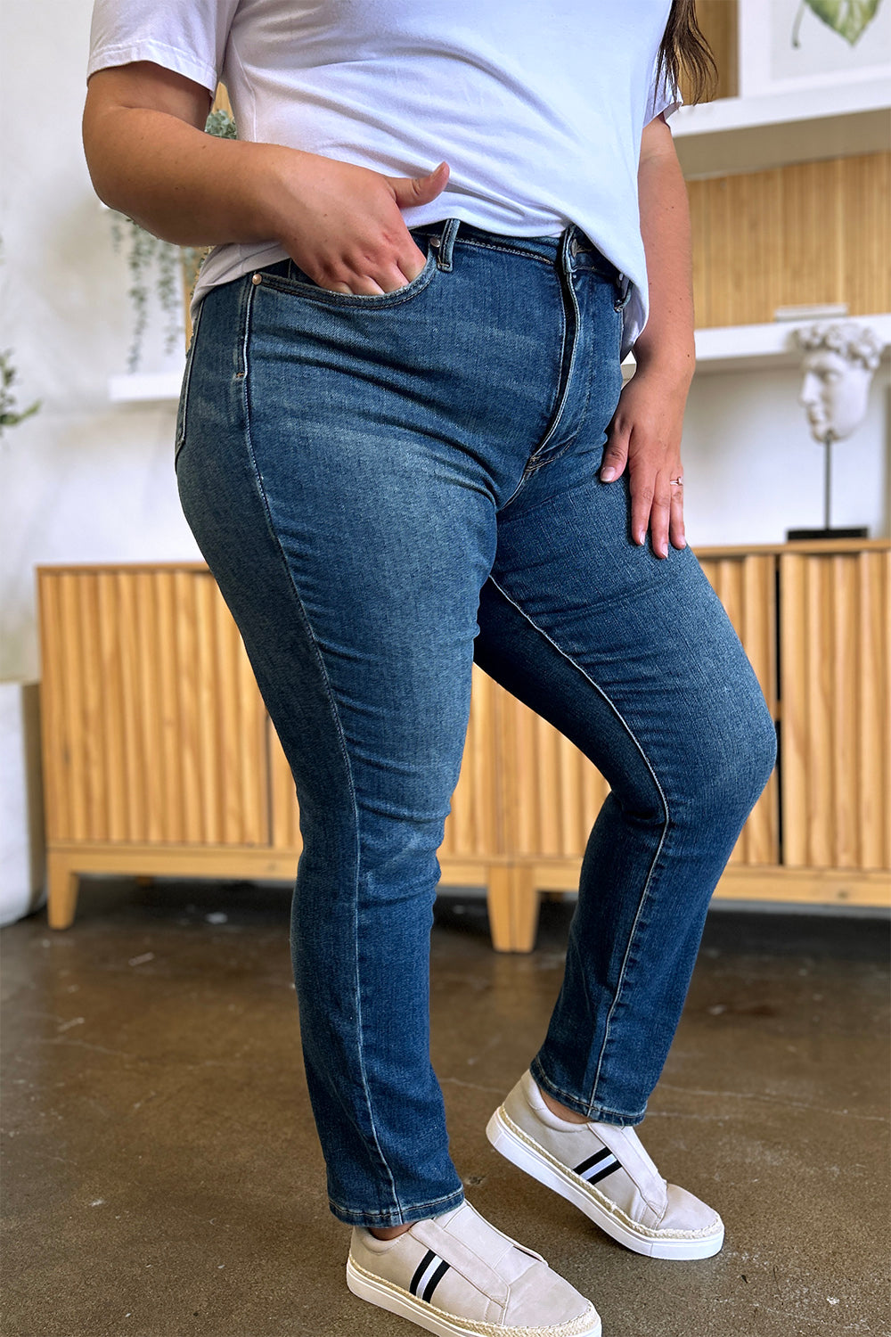 Person wearing Judy Blue Full Size Tummy Control High Waist Slim Jeans in blue and a white shirt, with their hands in their pockets. They are standing indoors on a polished concrete floor with a wooden cabinet and plant in the background.