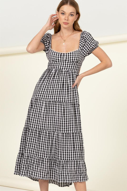 Wearing the "Somewhere to Go Tie-Back Gingham Print Maxi Dress," which features a black and white gingham print with puffed sleeves, a tiered skirt, and elegant tie-back detail, a person poses stylishly paired with black sandals.