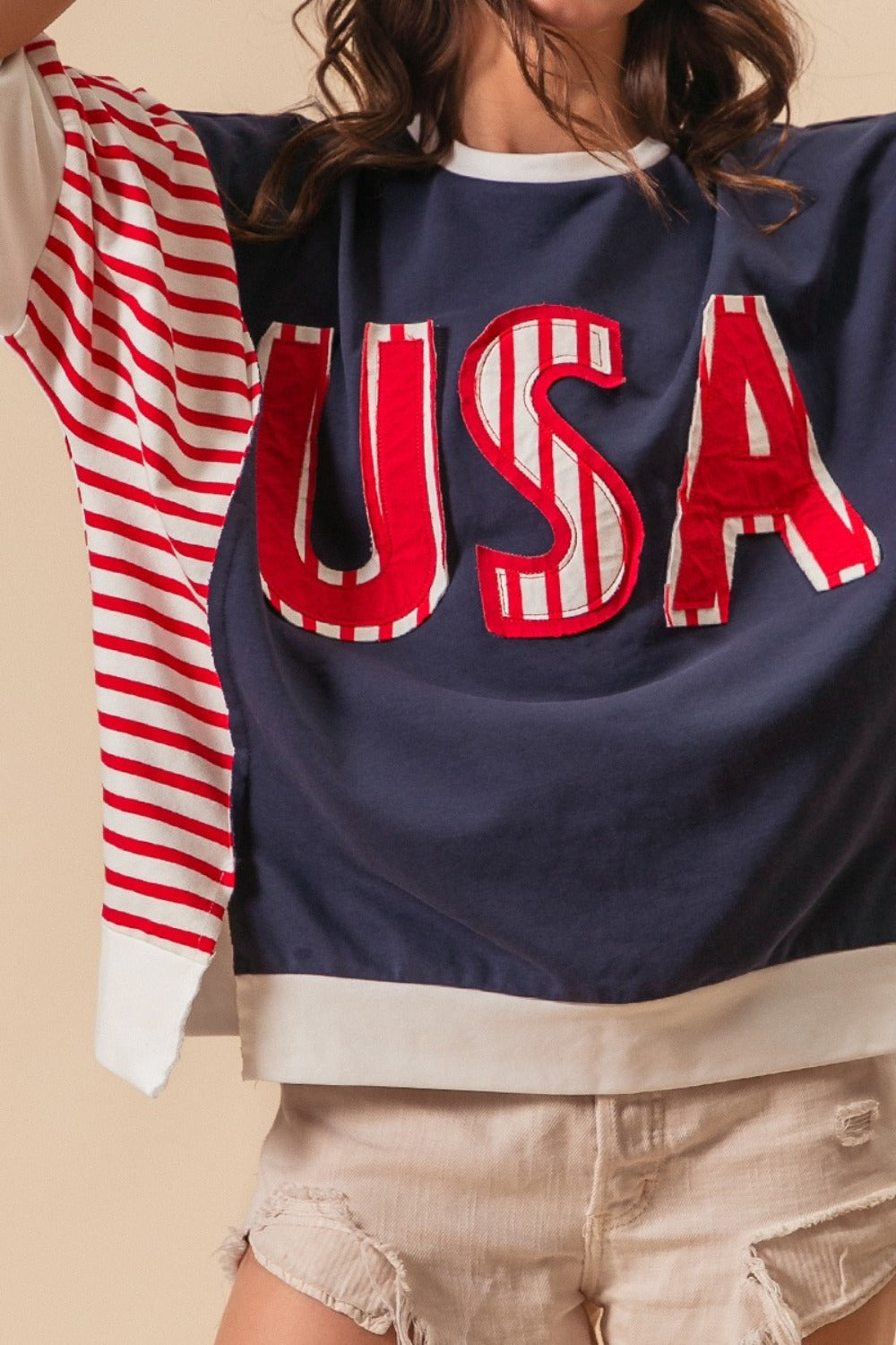 A person dressed in a “BiBi USA Letter Patchwork Contrast Short Sleeve T-Shirt” featuring blue, red, and white colors with striped sleeves, paired with beige shorts, poses against a beige background, proudly displaying their American spirit.