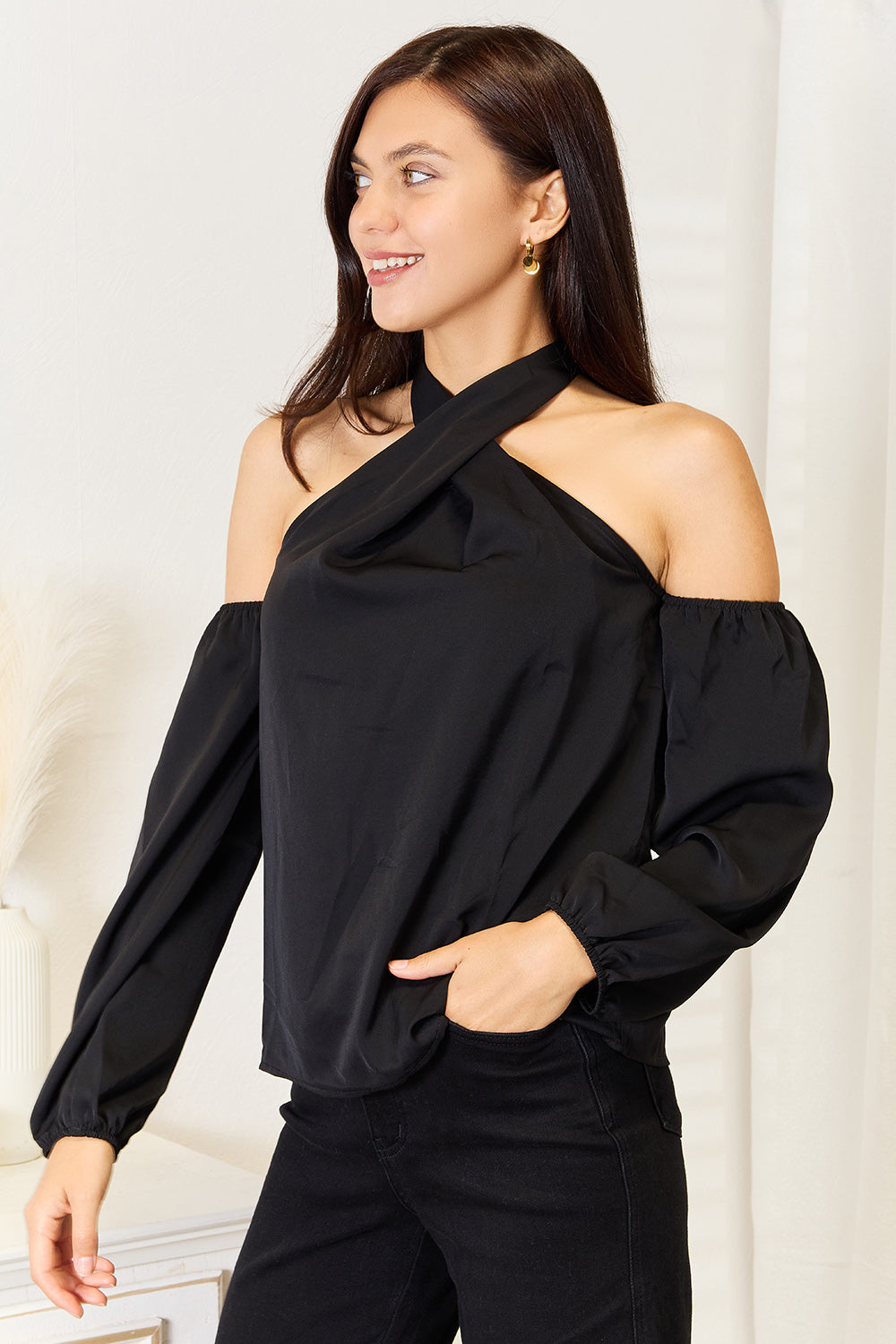 A woman with long brown hair wearing a Perfee Grecian Cold Shoulder Long Sleeve Blouse stands in a brightly lit room next to a white vase with pampas grass. She looks to the side with a smile.