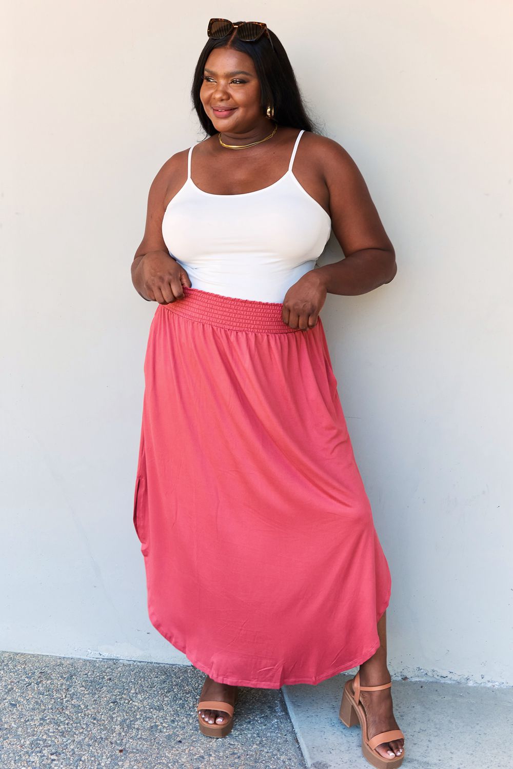 A person wearing a white top and the Doublju Comfort Princess Full Size High Waist Scoop Hem Maxi Skirt in an elegant coral color, paired with tan sandals, stands in front of a plain wall.