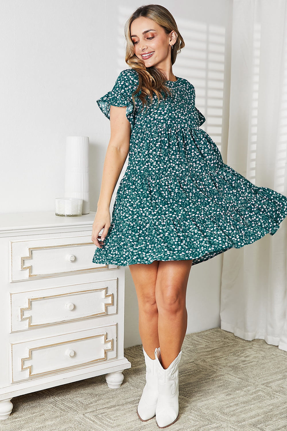 A woman wearing the Double Take Short Flounce Sleeve Tiered Dress in pink and red leopard print, complete with a ruffled hem and short flounce sleeves, smiles as she adjusts her brown hat. She stands near a plant, embodying the perfect boho-chic look.