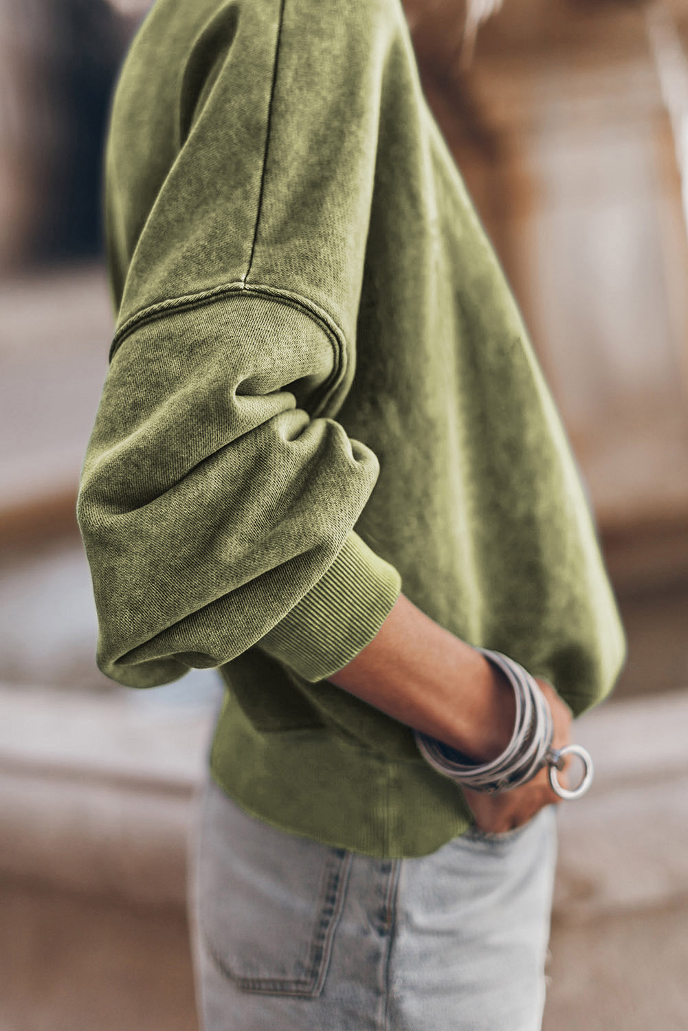 Person wearing a Green Drop Shoulder Crew Neck Pullover Sweatshirt with rolled-up sleeves, grey jeans, and bracelets. Their hand is in their pocket.